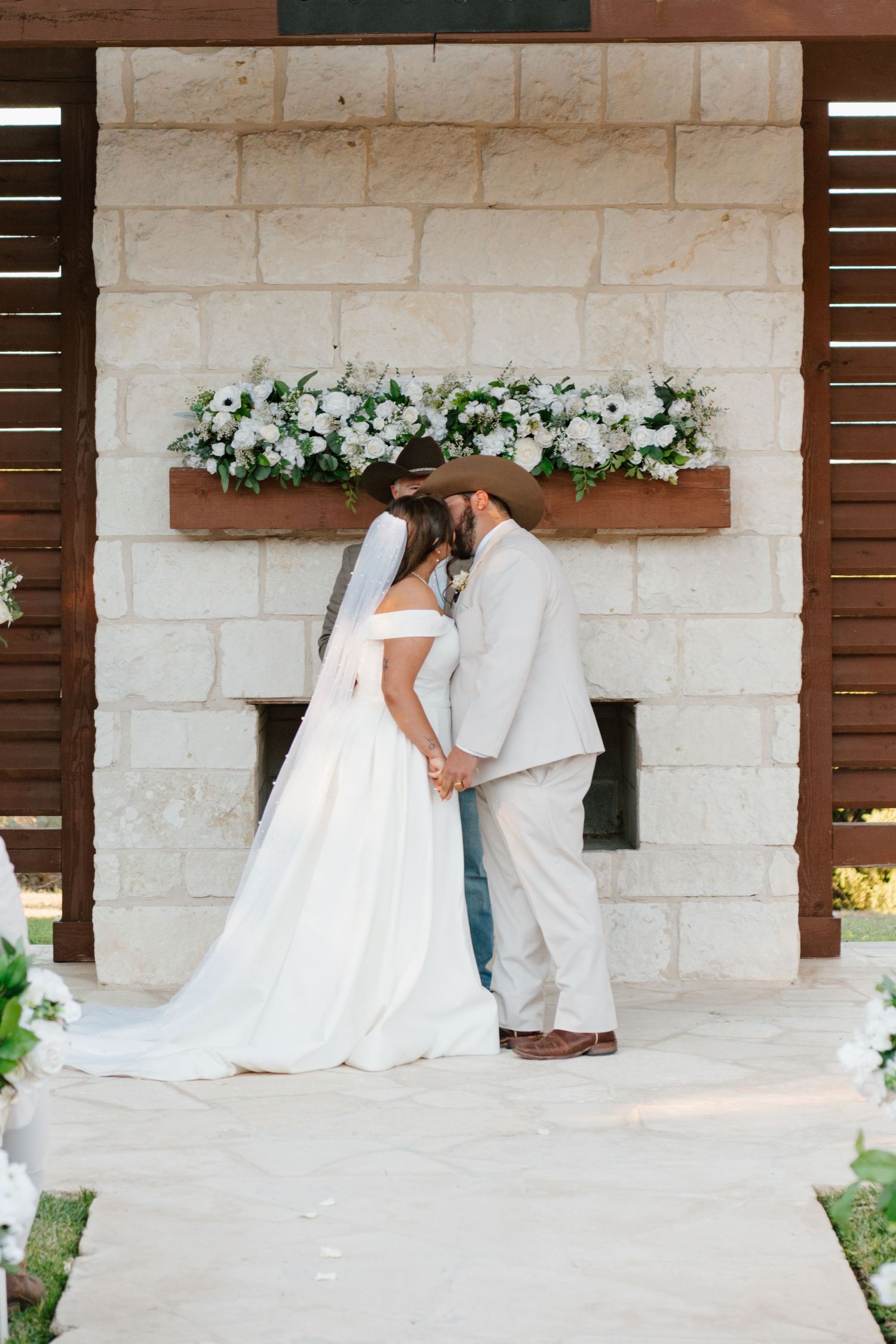 A couple shares a tender moment on their wedding day under a white stone archway adorned with white floral decorations.