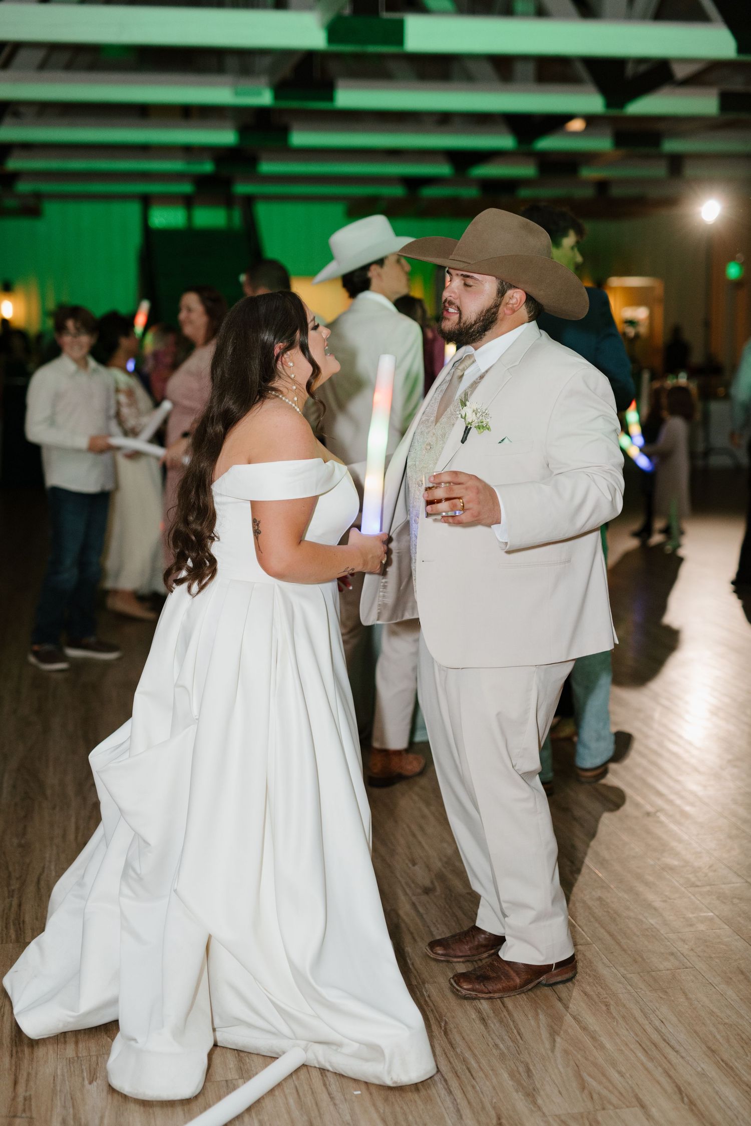 A couple in western-style wedding attire dances together on a wooden floor at their reception under green lighting.