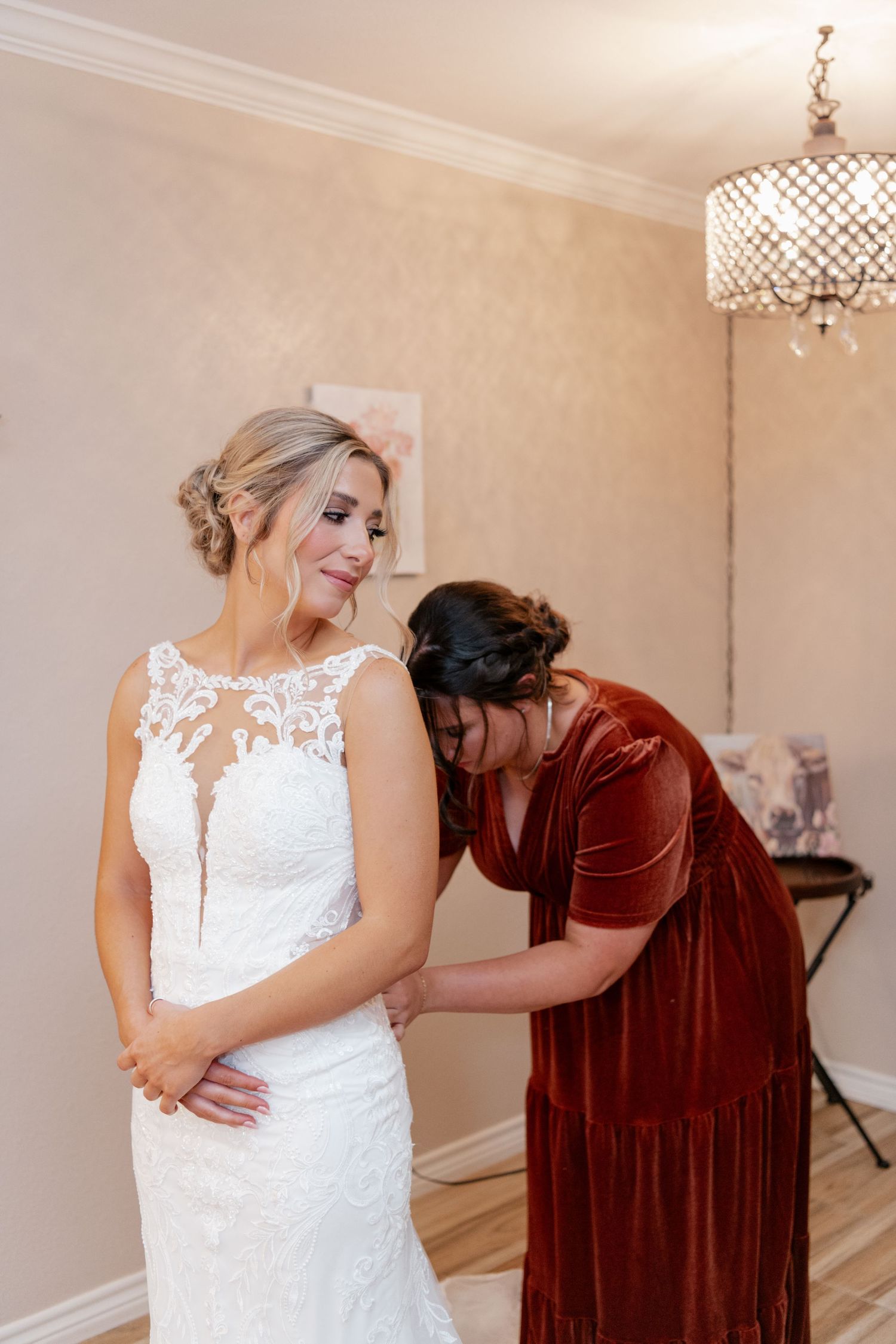 Wedding dress preparation scene with white lace gown being adjusted in elegant room with chandelier.