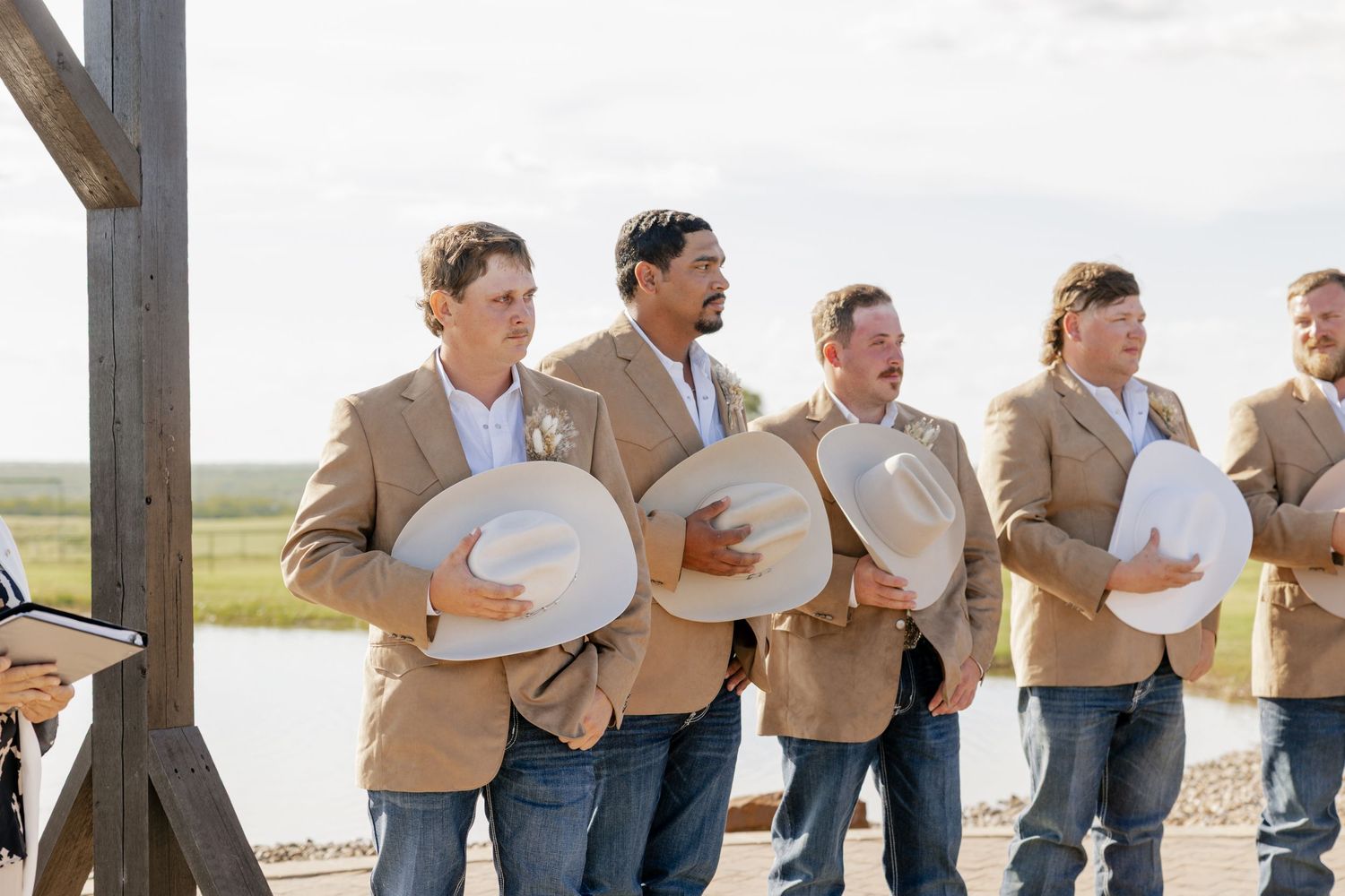 Groomsmen in tan jackets holding cowboy hats while standing by a waterfront.