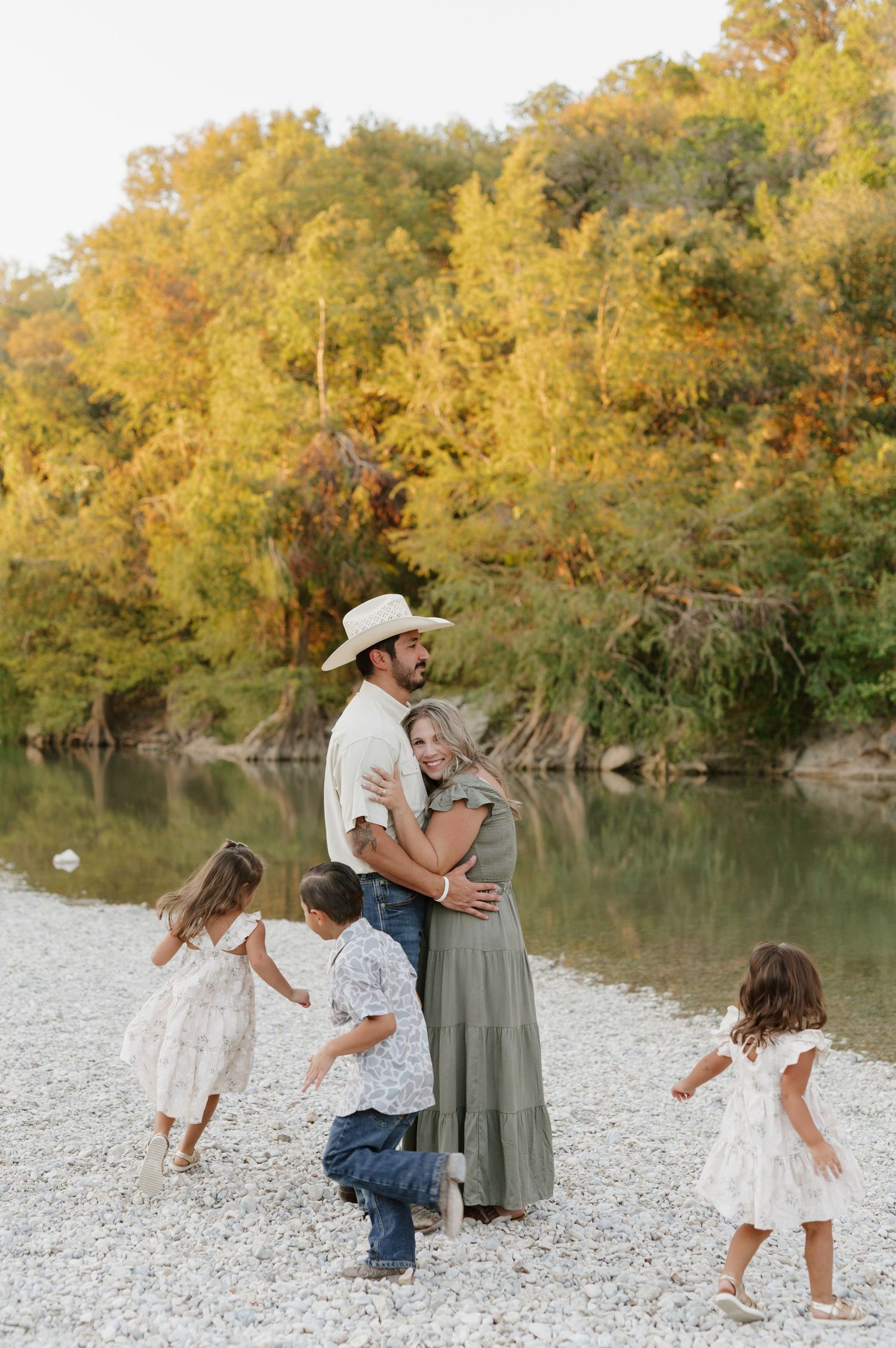 A family plays together on a rocky riverbank with golden autumn trees reflecting in the calm water during sunset.