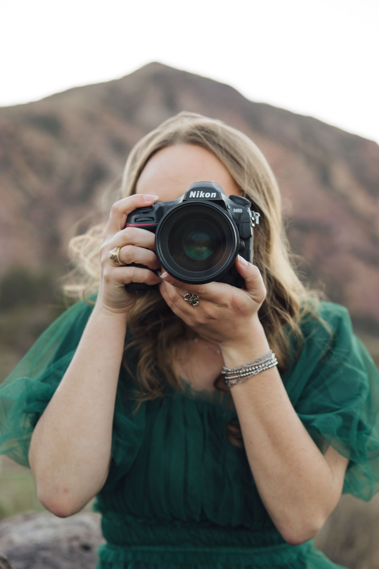 Photographer in emerald green dress holds Nikon DSLR camera against backdrop of desert mountains.