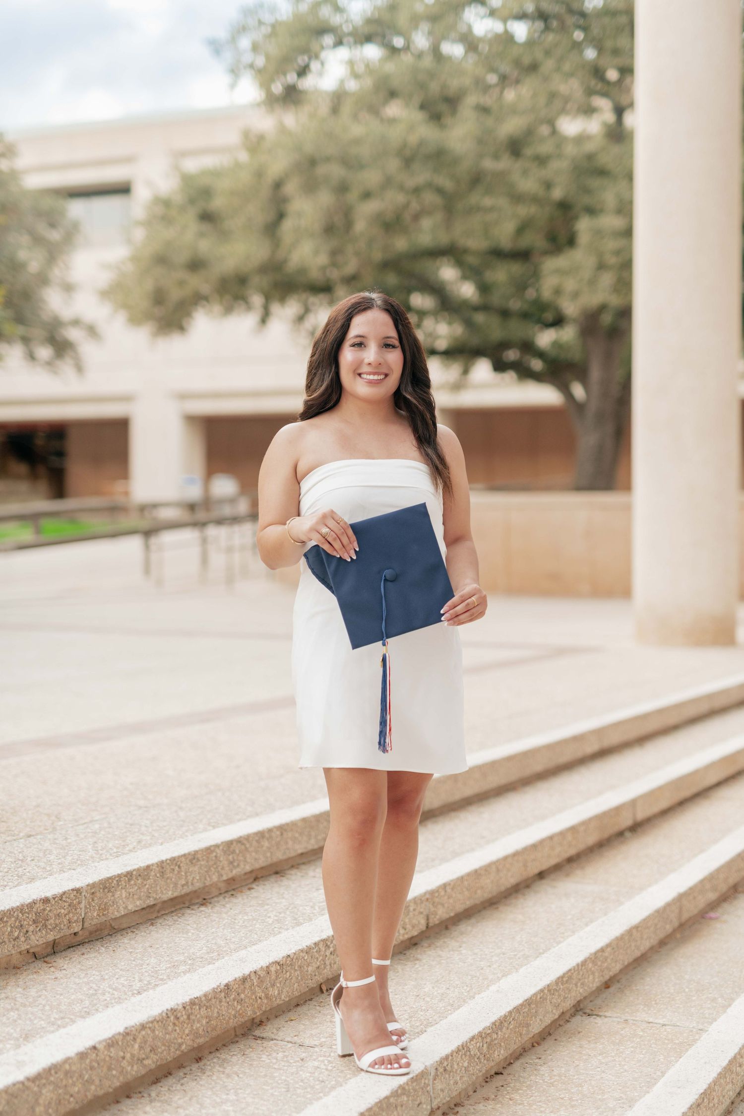 Graduate in white dress poses with diploma on campus steps.