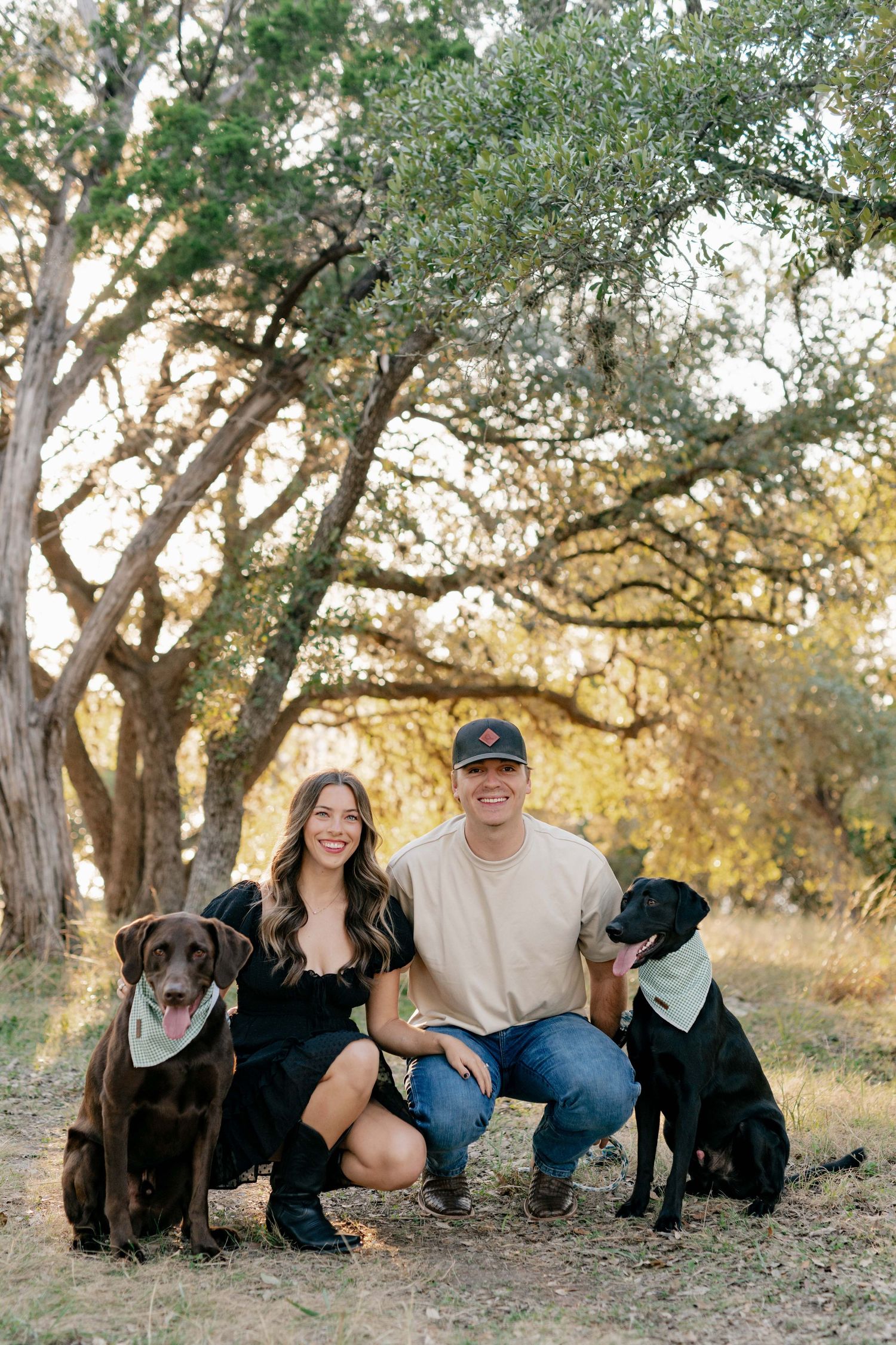 A happy couple kneels with their two dogs in a sun-dappled wooded setting.
