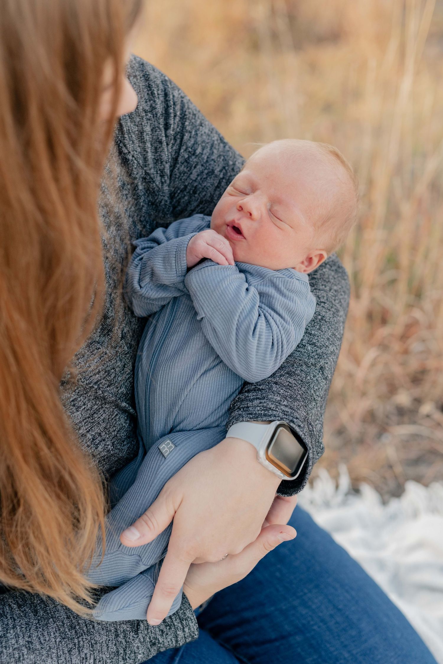 A sleeping newborn baby in a blue outfit being cradled in someone's arms wearing a gray sweater.