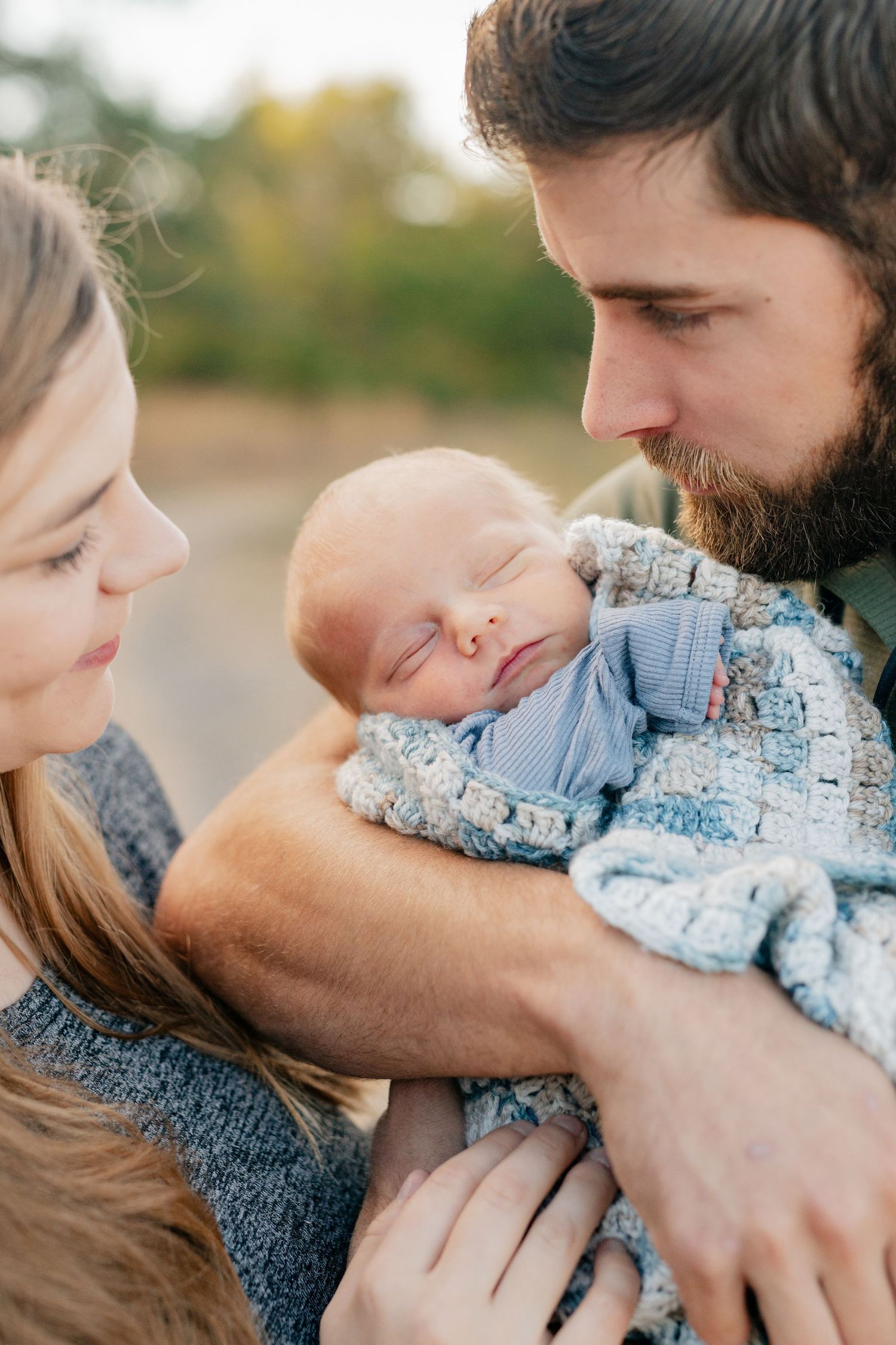Intimate close-up of parents cradling and kissing their peaceful sleeping newborn in soft blue clothing.
