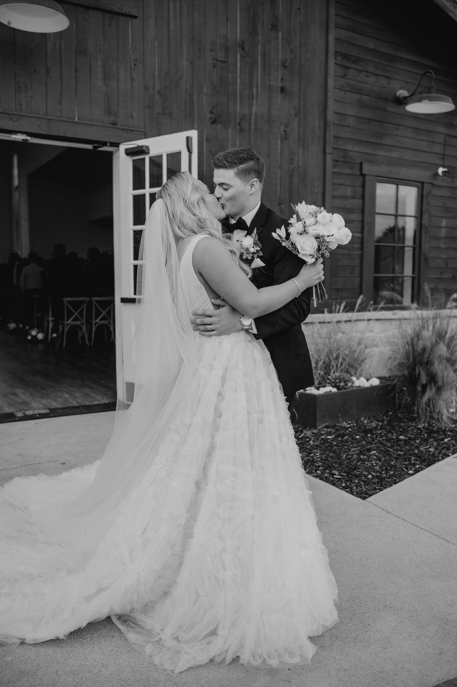 A bride and groom embracing and kissing on their wedding day in a rustic outdoor setting.