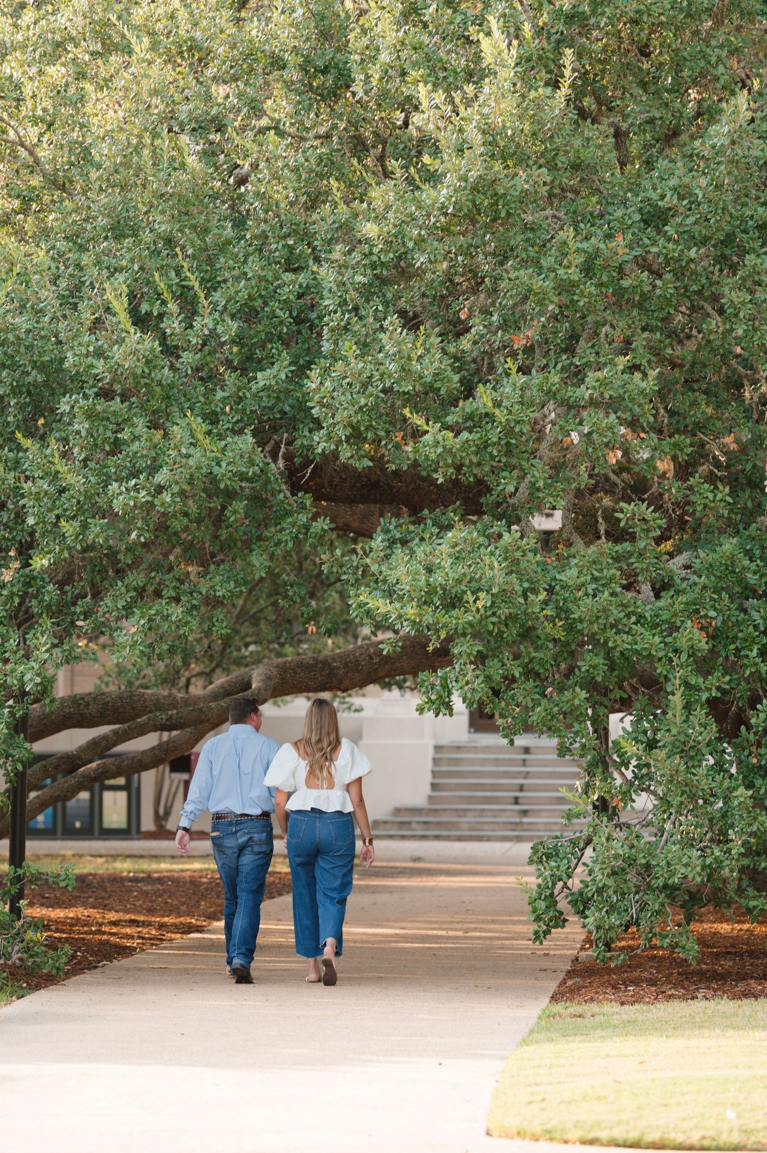 Two people in matching blue jeans walk away down a tree-lined path by concrete steps on a sunny day.