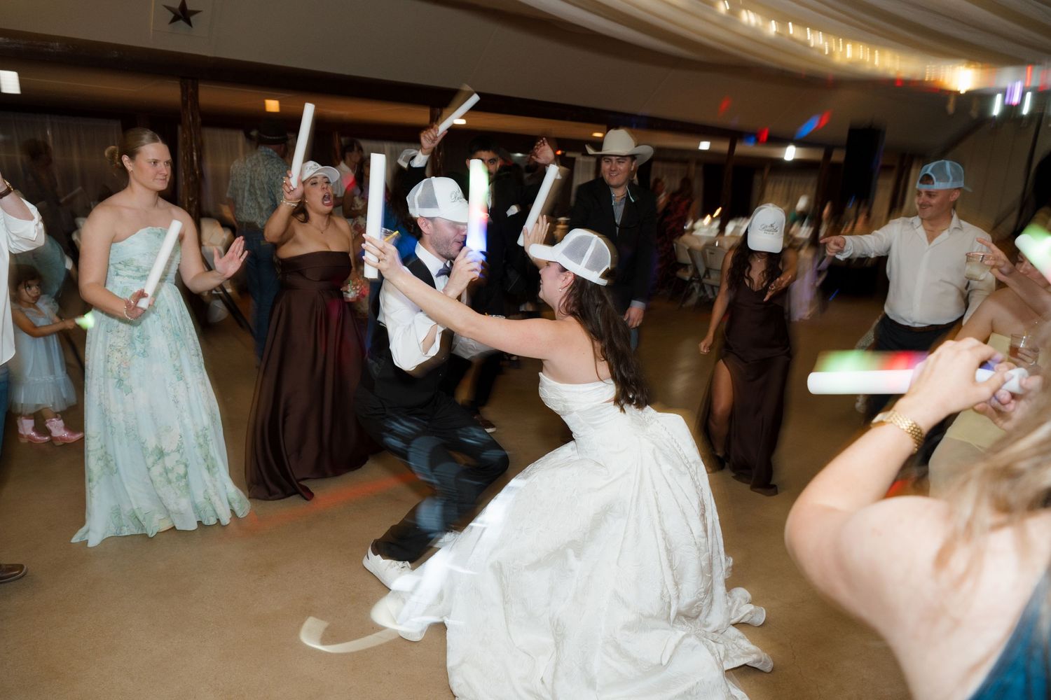 Bride and groom dance energetically with glowing batons surrounded by guests at lively indoor wedding reception.