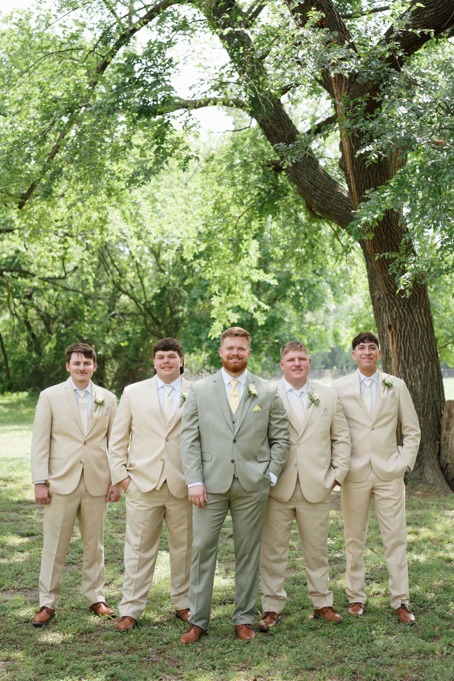 Groom and groomsmen pose in tan suits with boutonnieres under lush green trees outdoors.