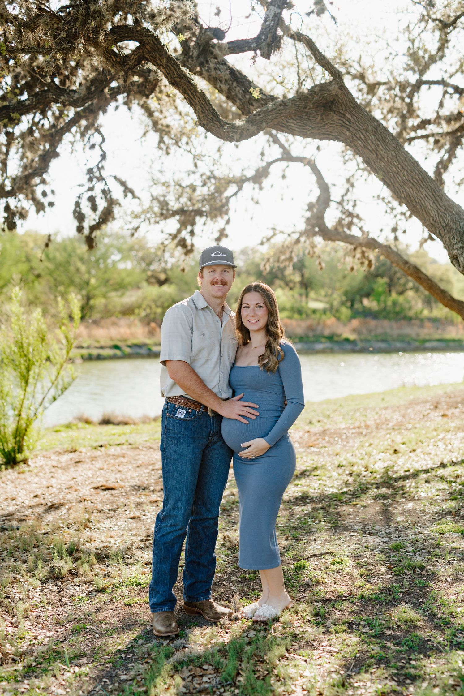 Expectant couple poses under a large oak tree by a serene lake during golden hour.