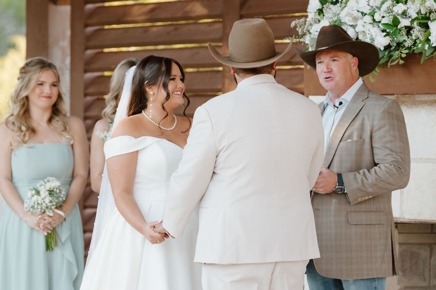A wedding ceremony takes place under a rustic wooden pergola with white floral decorations and mint green accents.