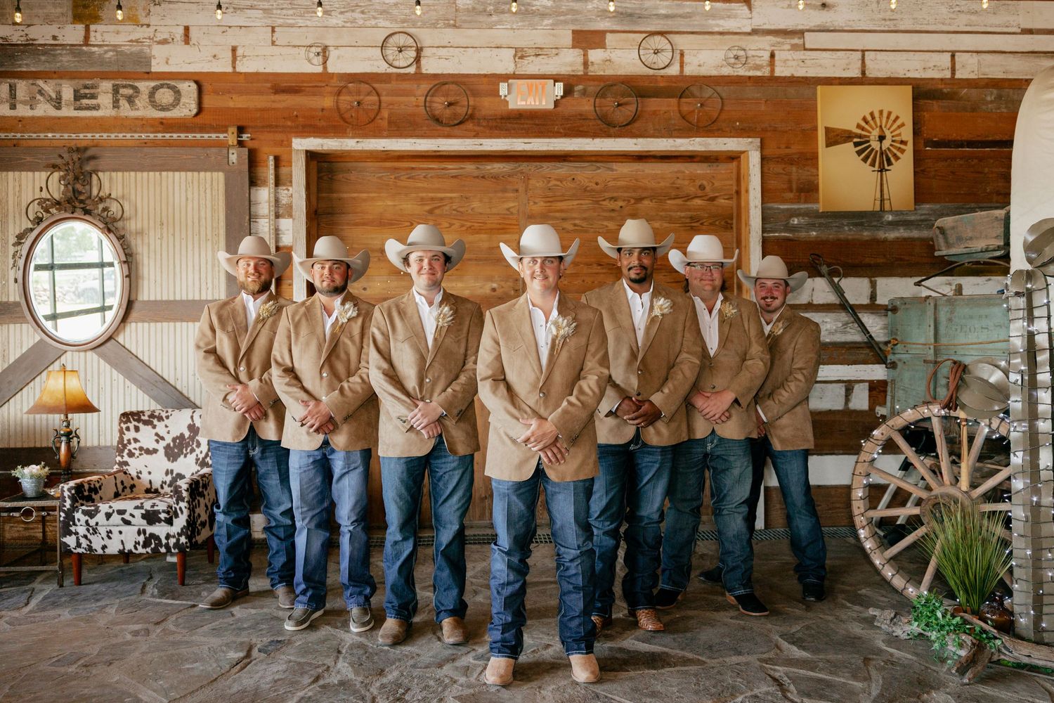 A group of cowboys in matching tan jackets and jeans lined up against a rustic wooden wall.