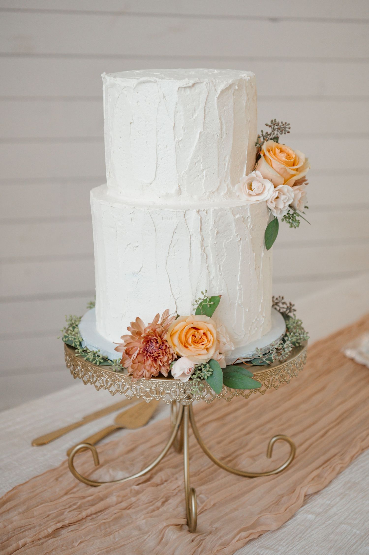 White wedding cake with peach roses and greenery displayed on an elegant gold stand against rustic wood background.