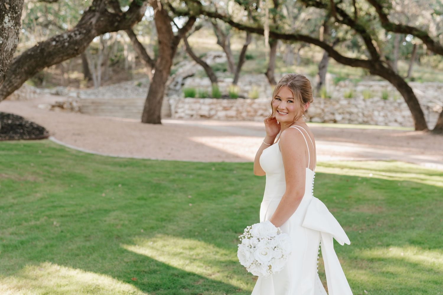 A person in a white lace dress poses in a sunlit garden setting with trees and grass in the background.