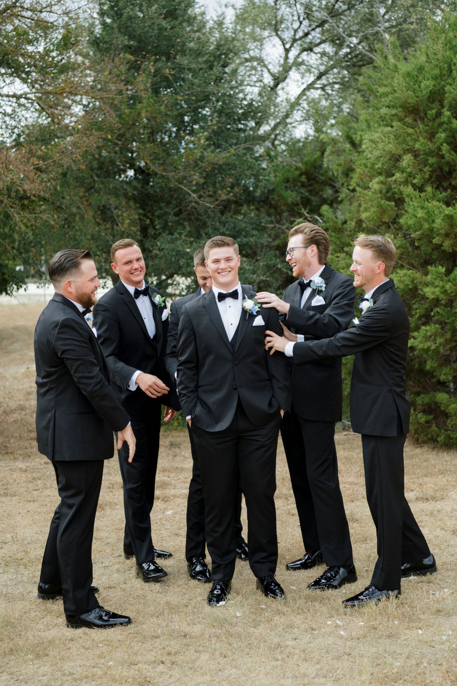 A group of men in formal attire, including suits and bow ties, standing together in an outdoor setting with trees in the background.