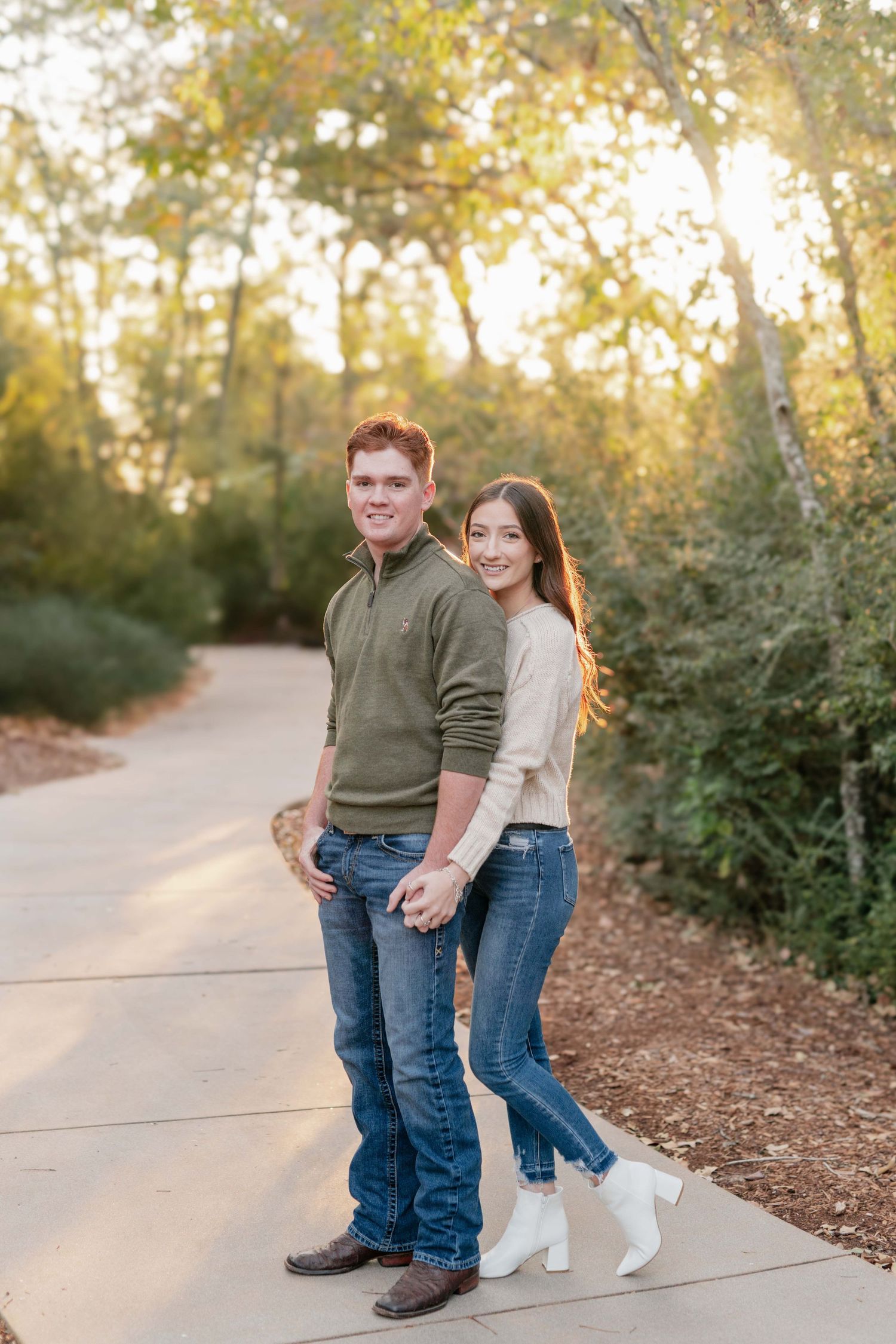 A couple stands together on a sunlit path surrounded by trees and autumn foliage.