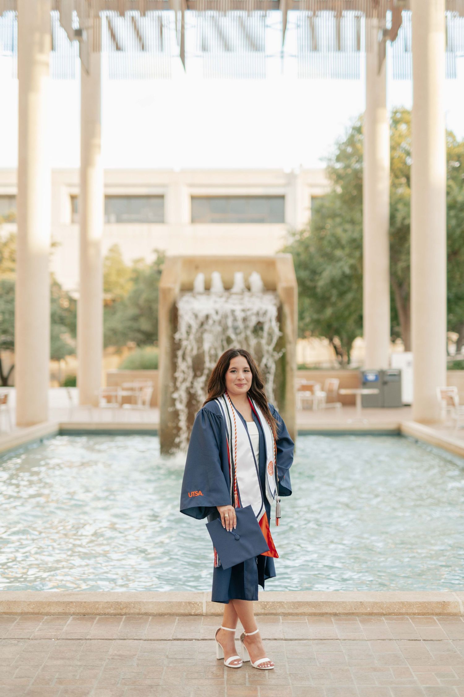 Graduate in academic regalia stands by campus fountain.