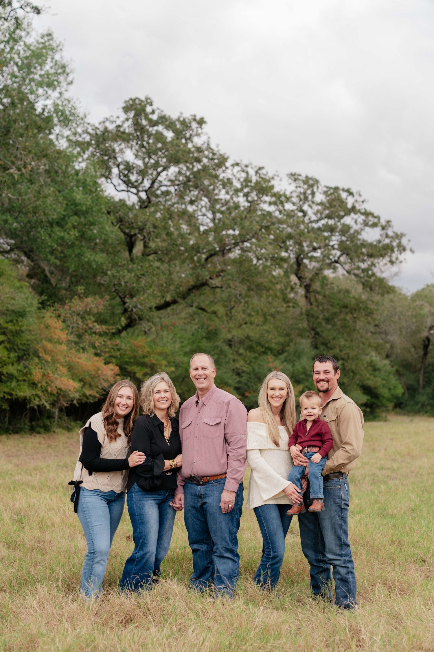 A family of six poses together in casual attire against a backdrop of oak trees in a grassy field.