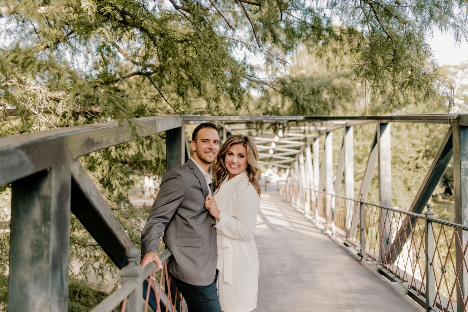Couple in elegant attire pose together on a rustic wooden footbridge surrounded by green foliage.