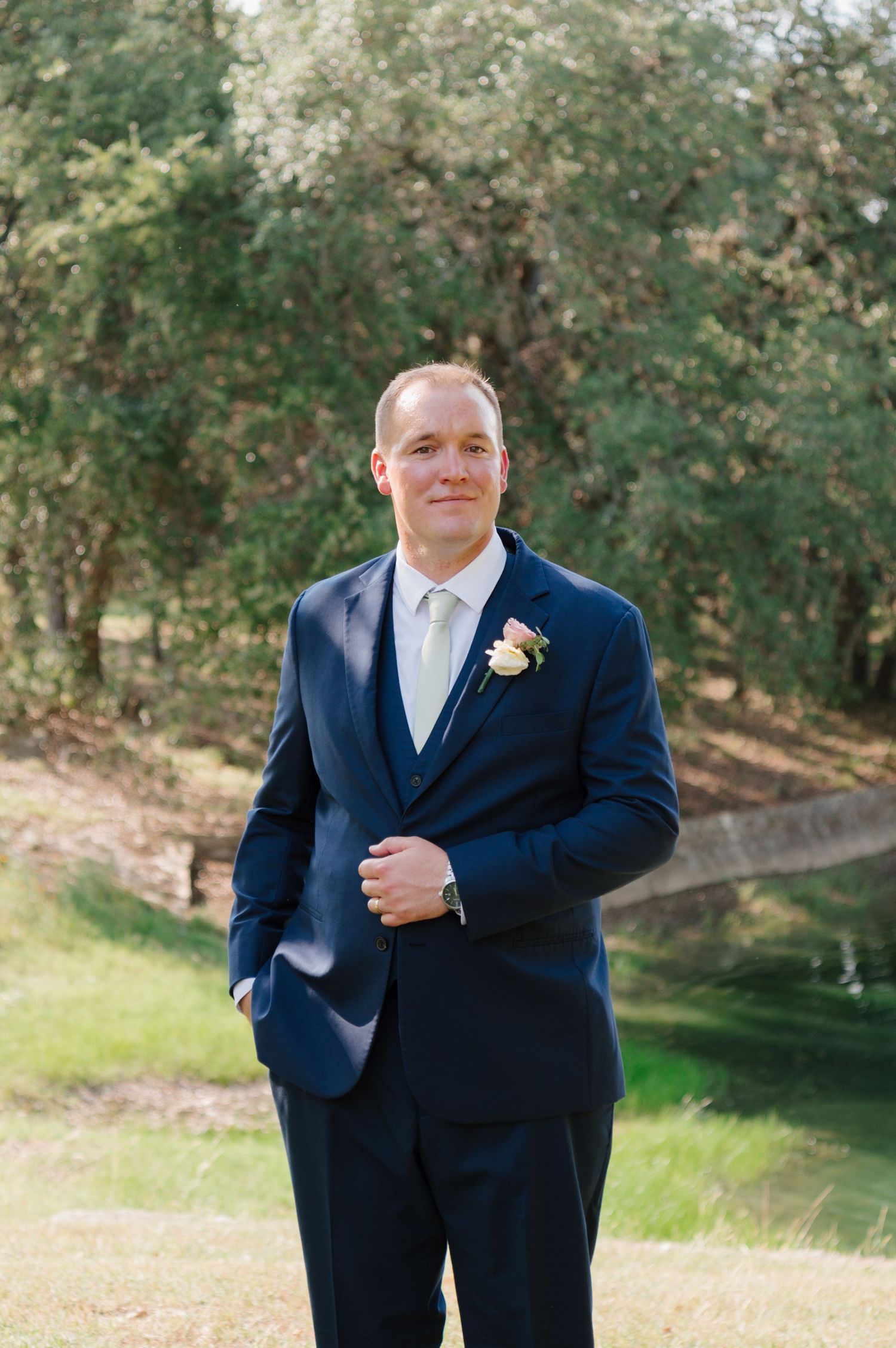 Groom in navy suit and cream tie poses outdoors on grassy hillside.