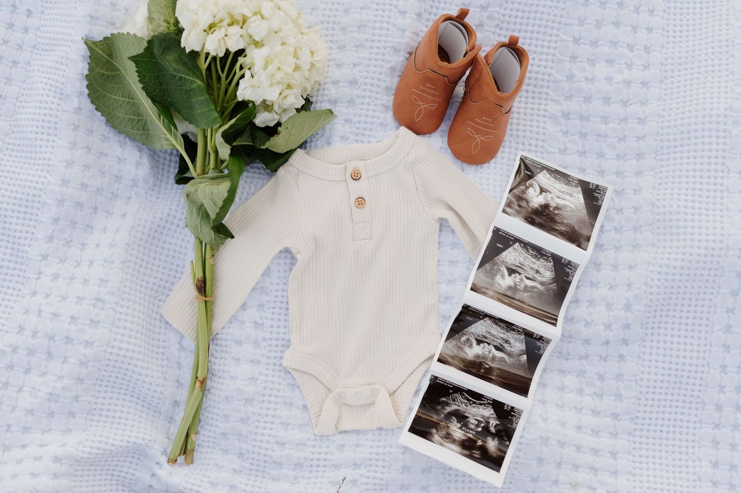 Flat lay of baby items including white onesie, flowers, and ultrasound photos on textured background.