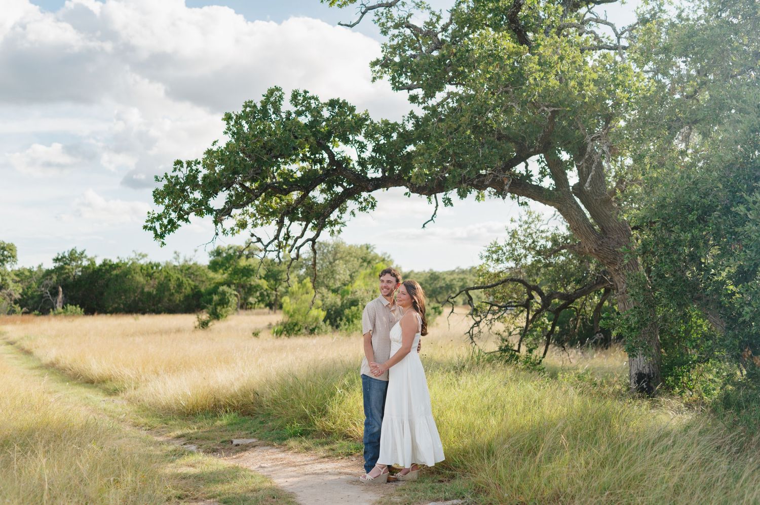 Two people stand together in a sunlit meadow with tall golden grass and green trees in the background.