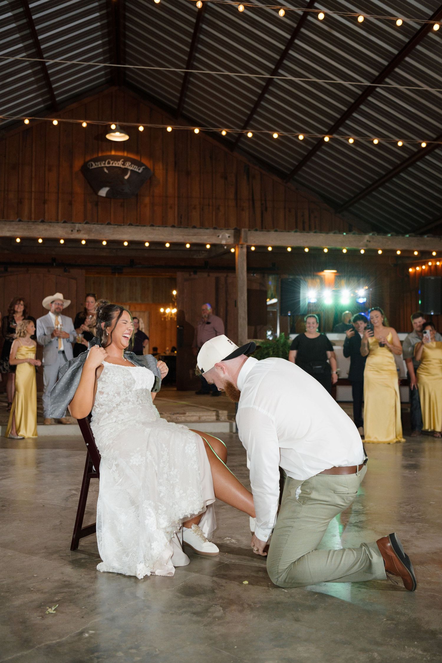 Groom kneels to remove bride's garter during lively barn wedding reception with string lights and guests in yellow dresses.
