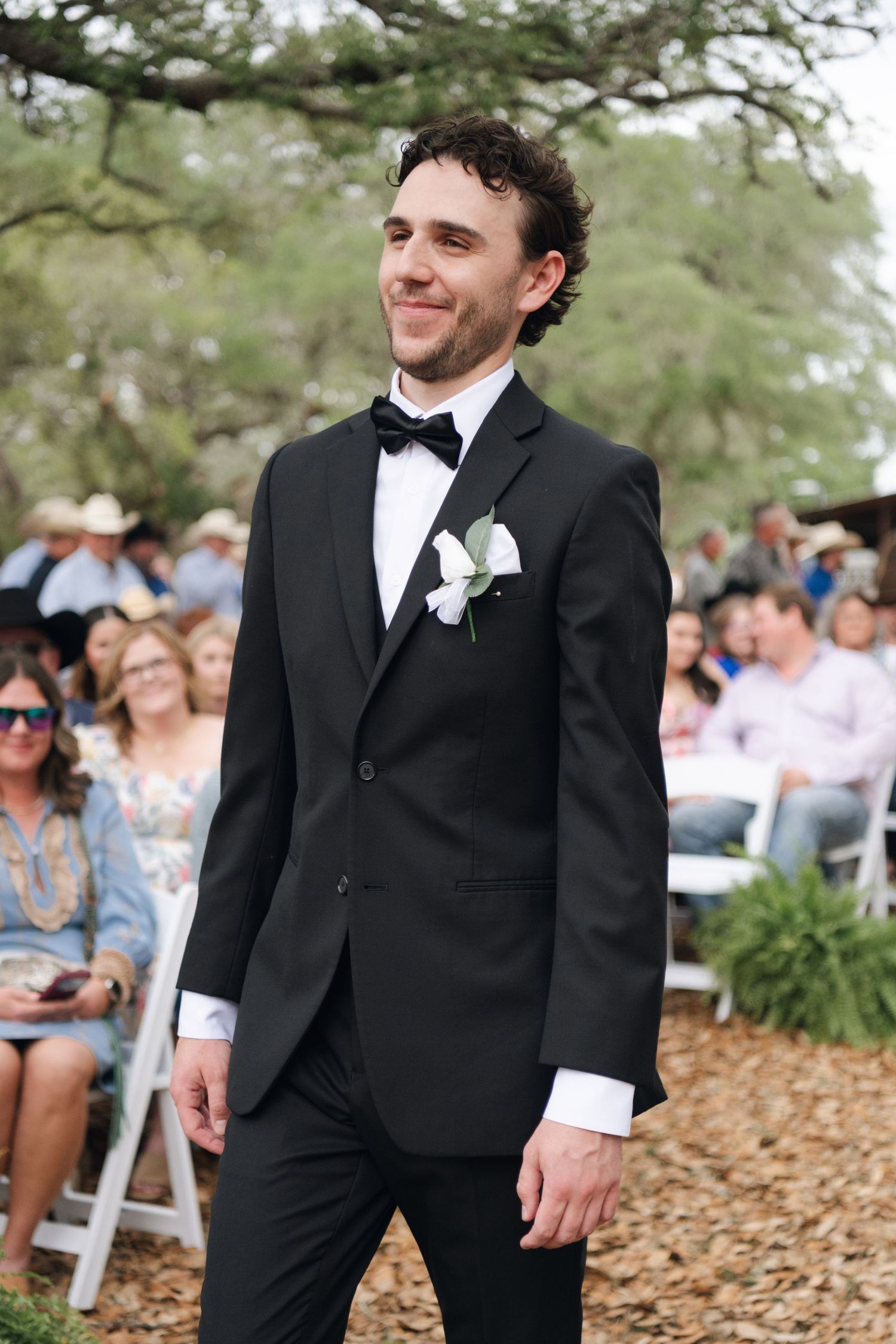 Dapper groom in black tuxedo with boutonniere smiles while walking down outdoor wedding aisle.