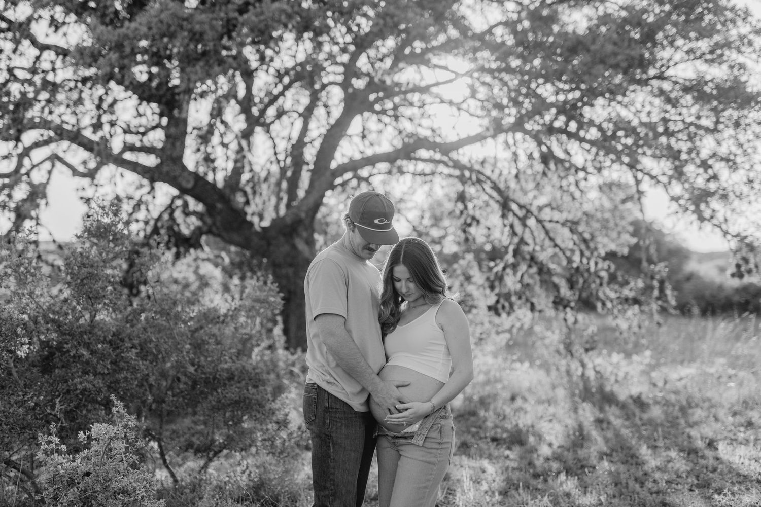 A couple embraces in a sun-dappled outdoor setting with trees in the background, captured in black and white.