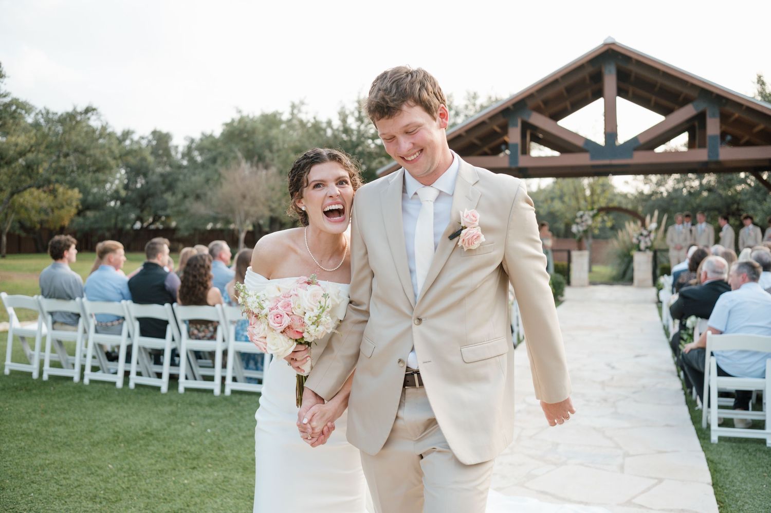 A romantic outdoor wedding ceremony setup with white chairs and a rustic wooden pergola in a garden setting.