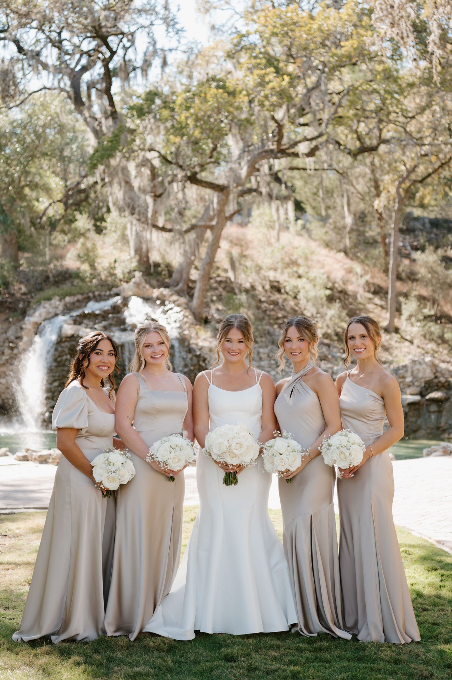 Bridal party in neutral champagne dresses poses outdoors under oak trees.