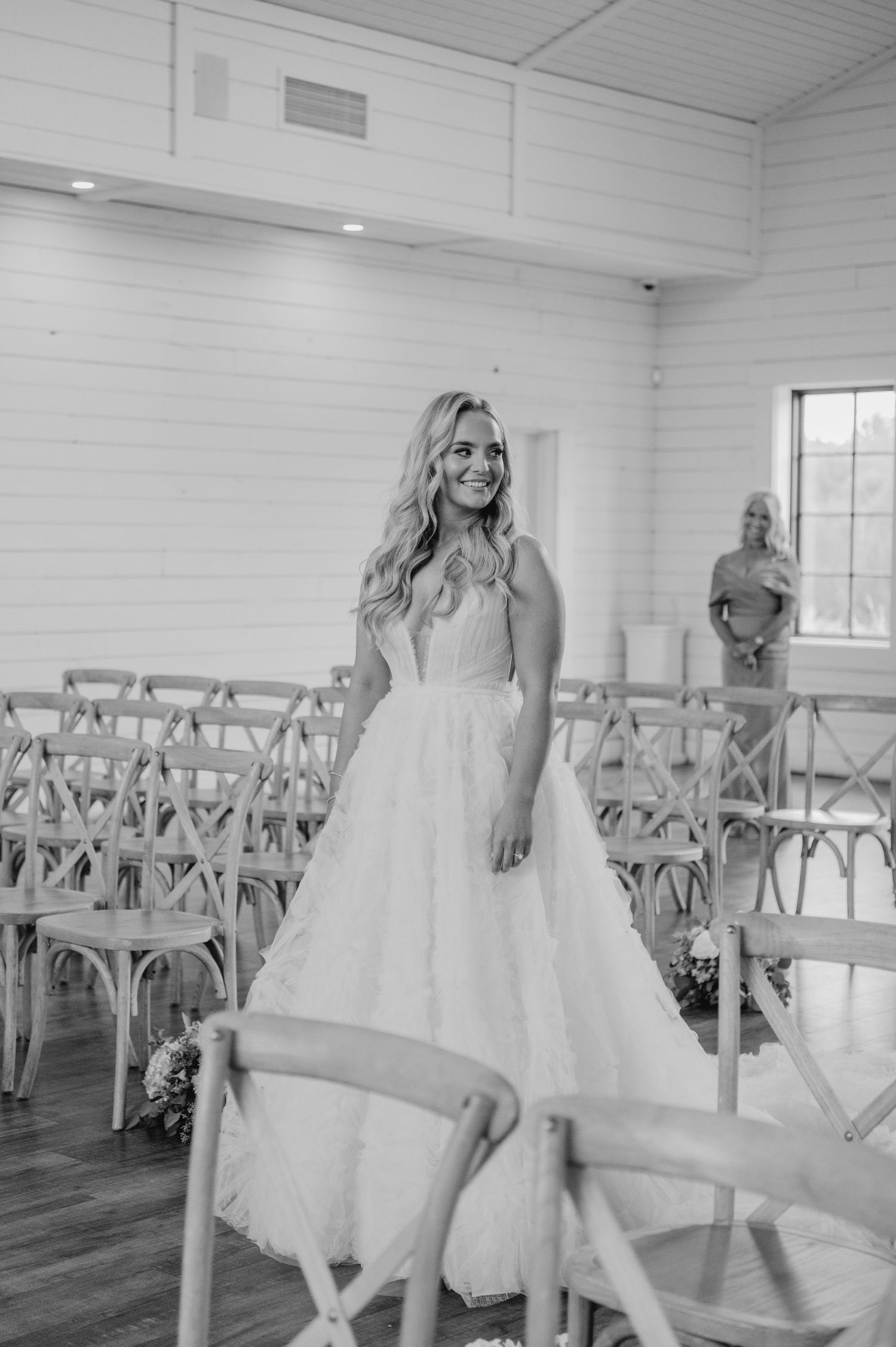A bride in a white wedding dress standing in a rustic indoor setting, with wooden walls and chairs in the background.