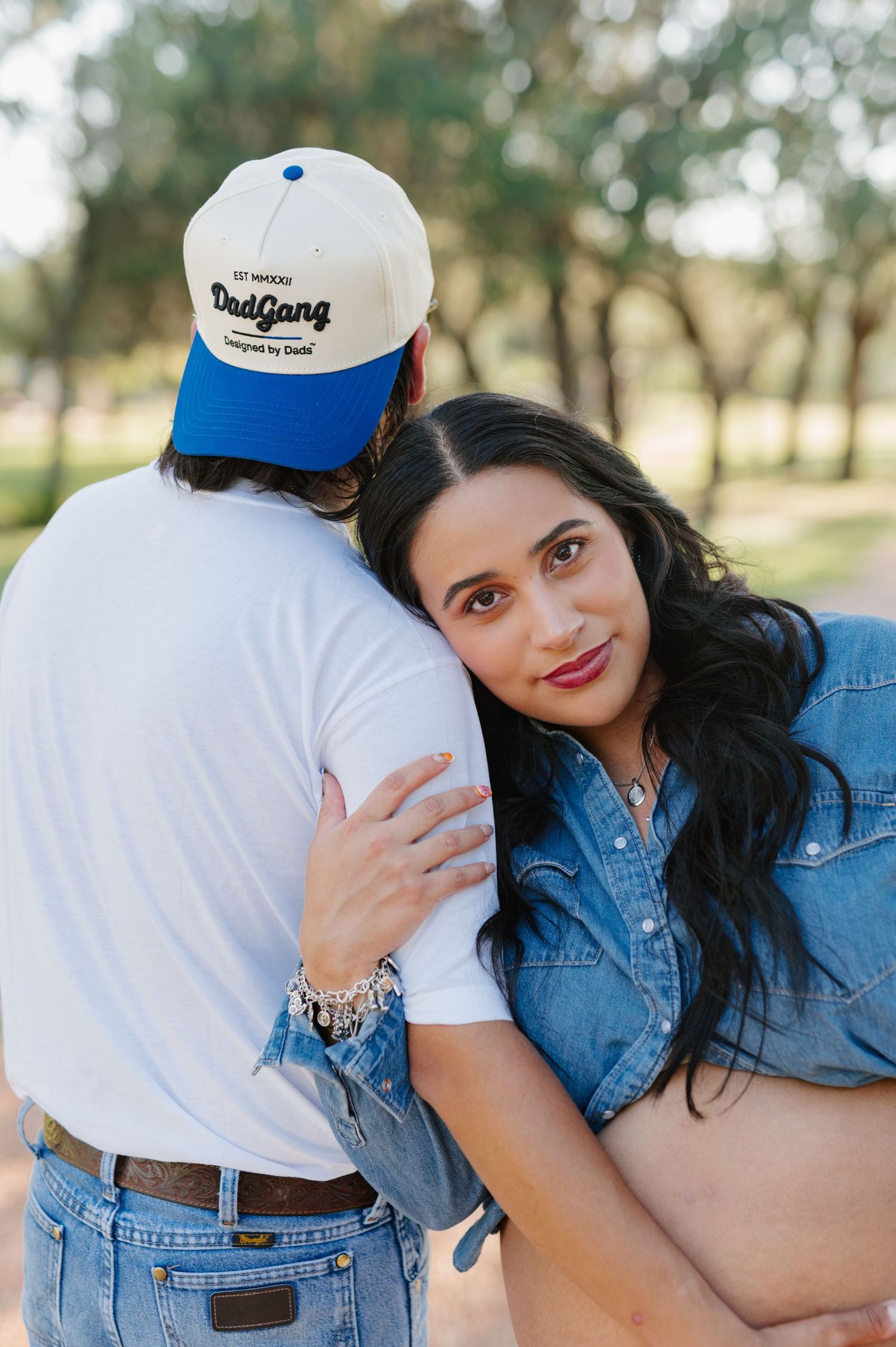 A couple poses together, with one wearing a denim jacket and the other in white with a baseball cap.