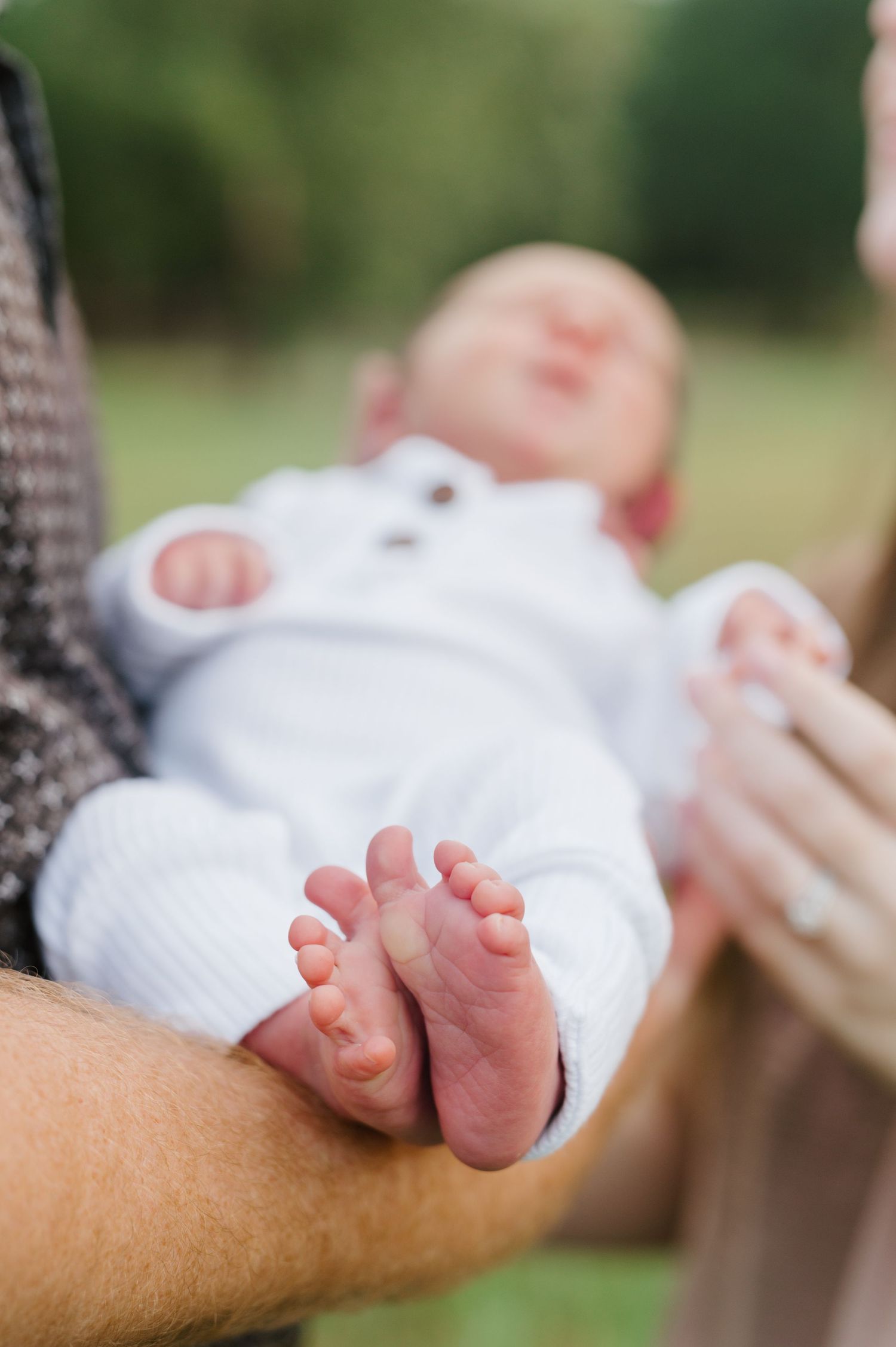 Small baby feet and toes peeking out from white blanket while resting on someone's arm in outdoor setting.