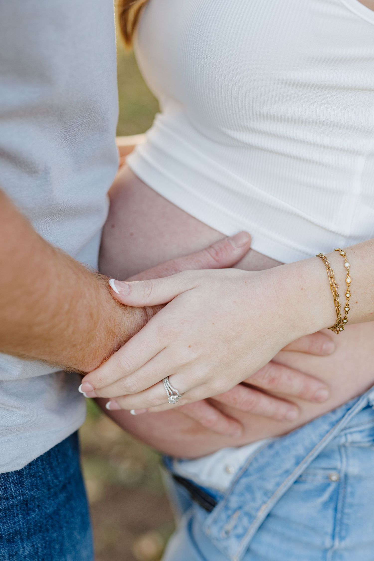 Close up of hands resting on a pregnant belly, showing a delicate gold bracelet and engagement ring.