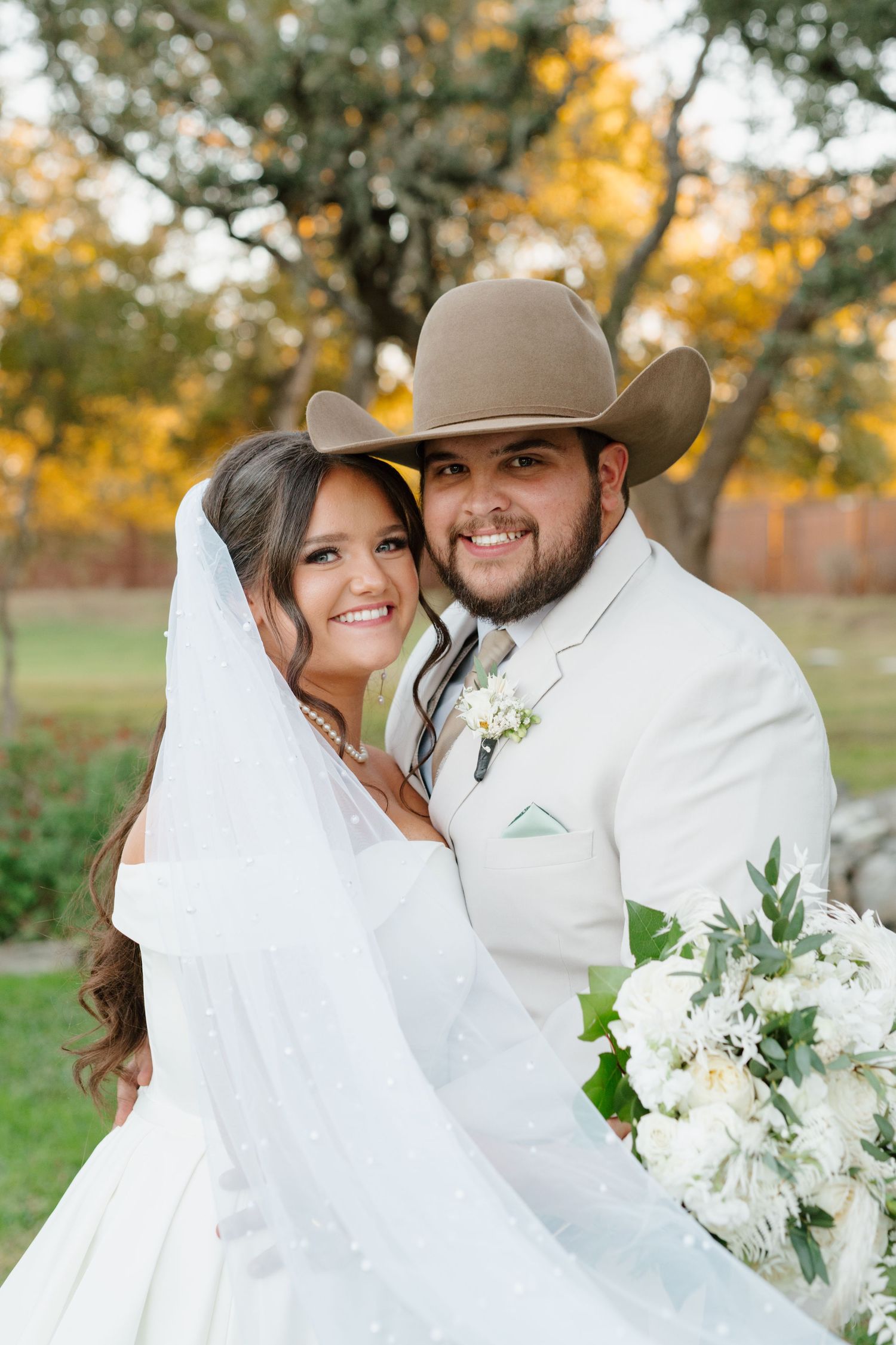 A rustic outdoor wedding portrait with the couple in white attire, featuring a cowboy hat and bridal bouquet in autumn setting.