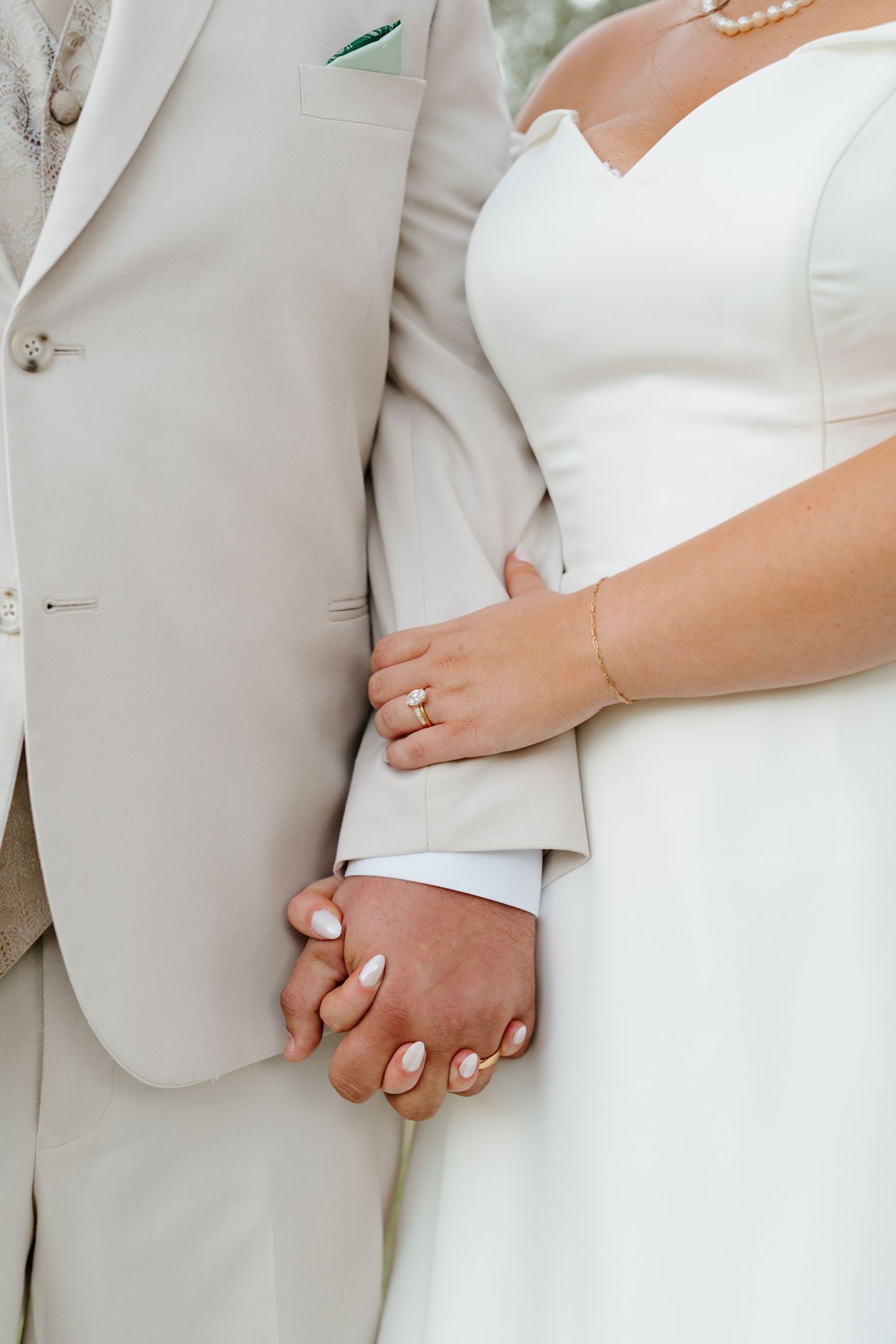 Close-up of newlywed couple holding hands, with bride in white dress and groom in light beige suit during wedding ceremony.