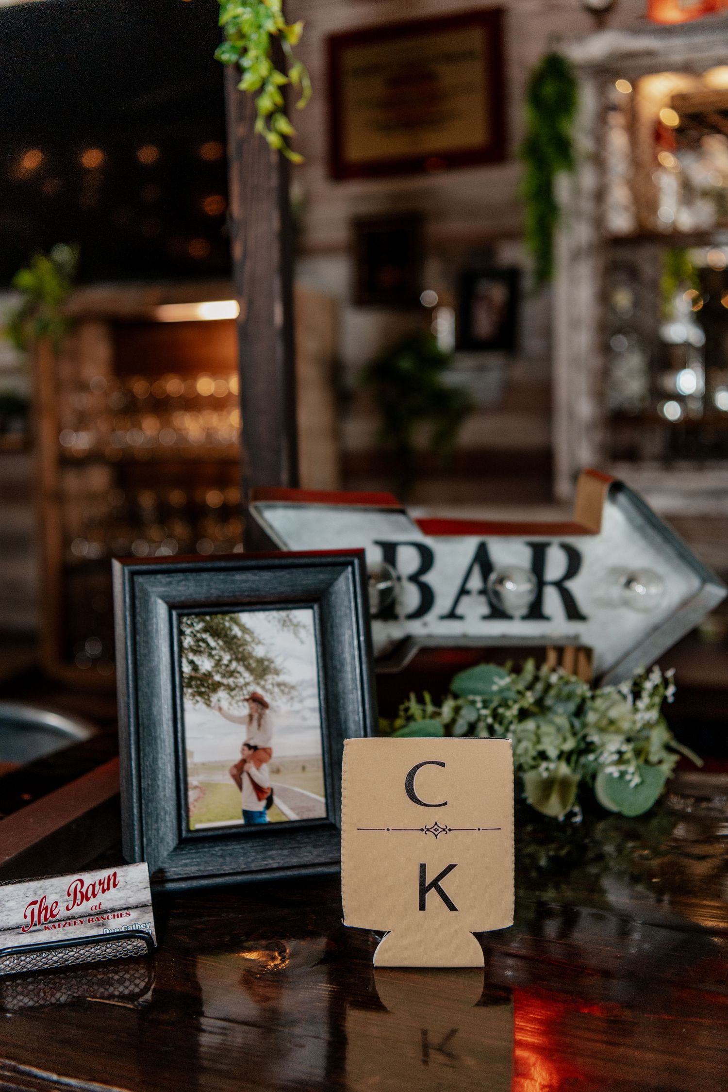 Rustic bar decor featuring a framed photo, monogrammed drink koozie, and vintage arrow sign in warm ambient lighting.