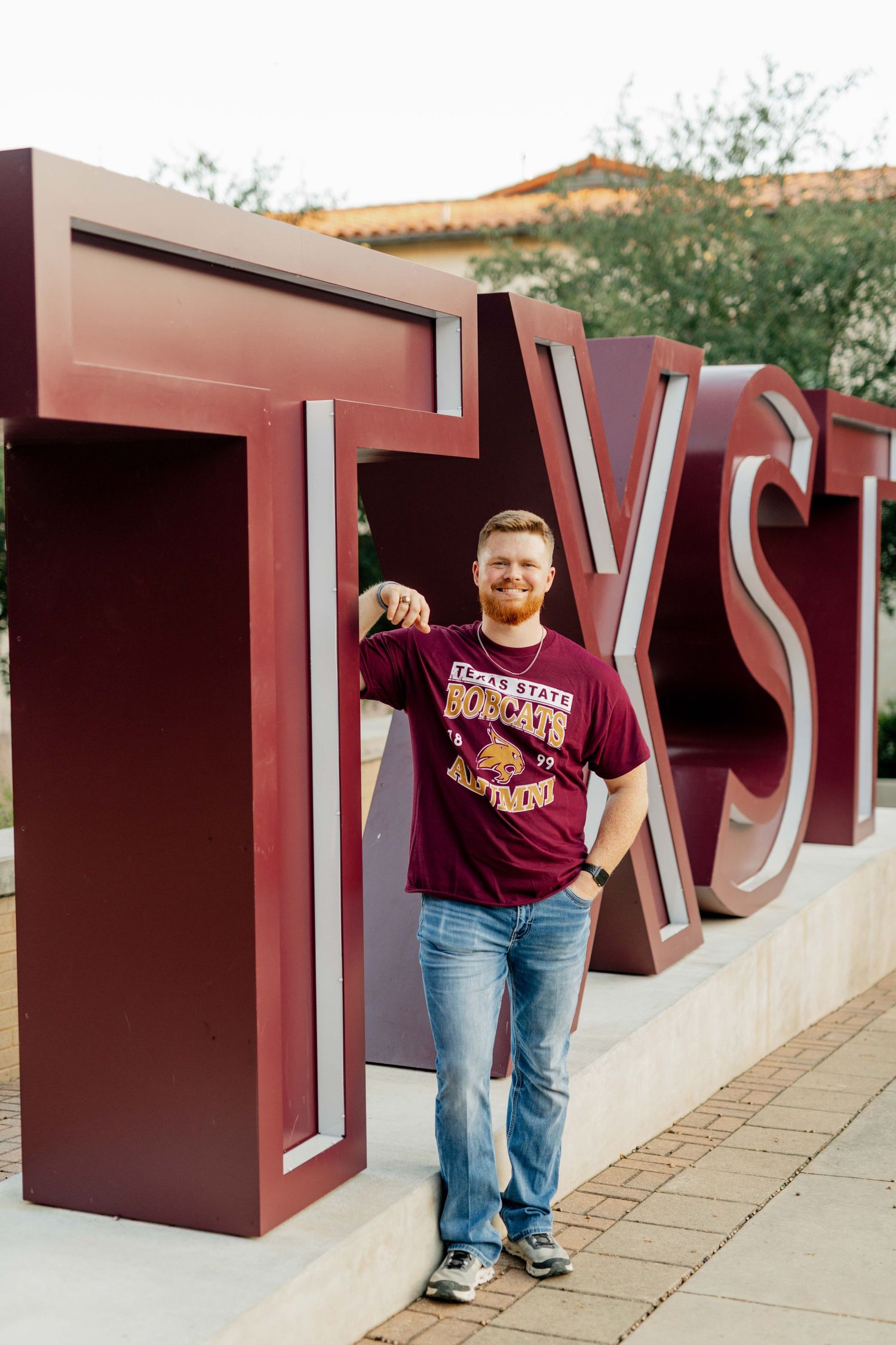 Graduate poses near TXST letters on Texas State University's campus during sunset wearing a TXST t-shirt.