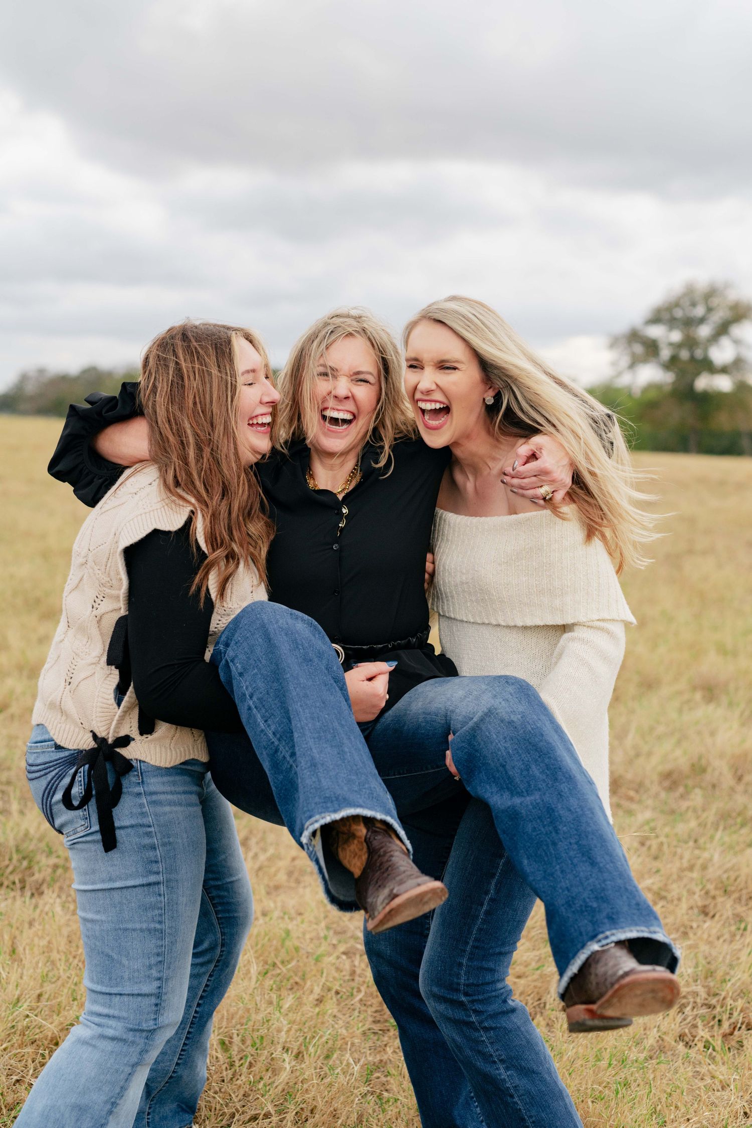 Three friends share joyful laughter while playfully carrying each other in a sunlit field.