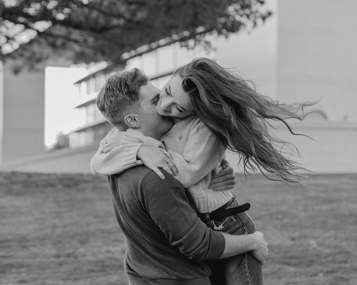 A romantic black and white photo of a couple embracing with windblown hair on a hill.