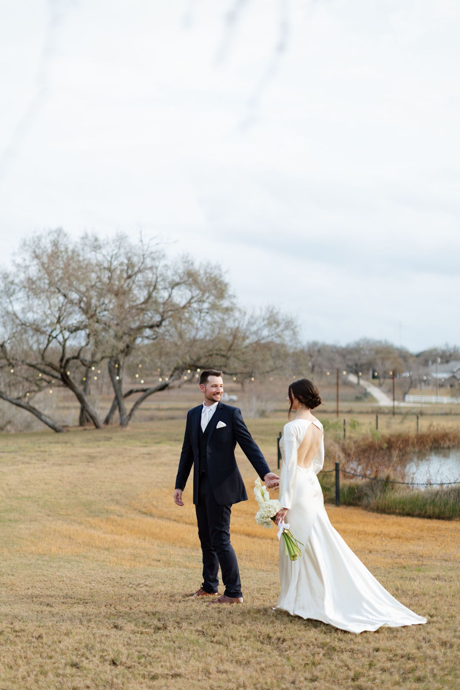 A couple stands together on a grassy field with bare trees and water in the background on their wedding day.