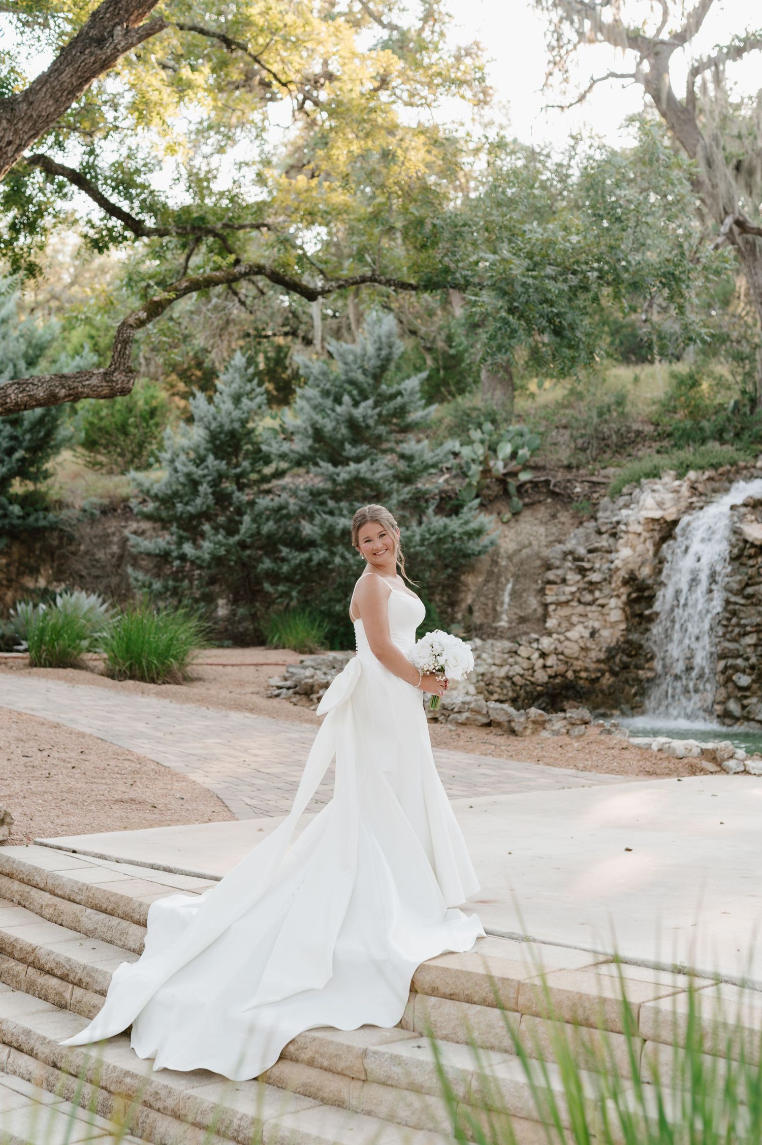 Bride in elegant white dress with long train poses by stone wall and waterfall in garden.