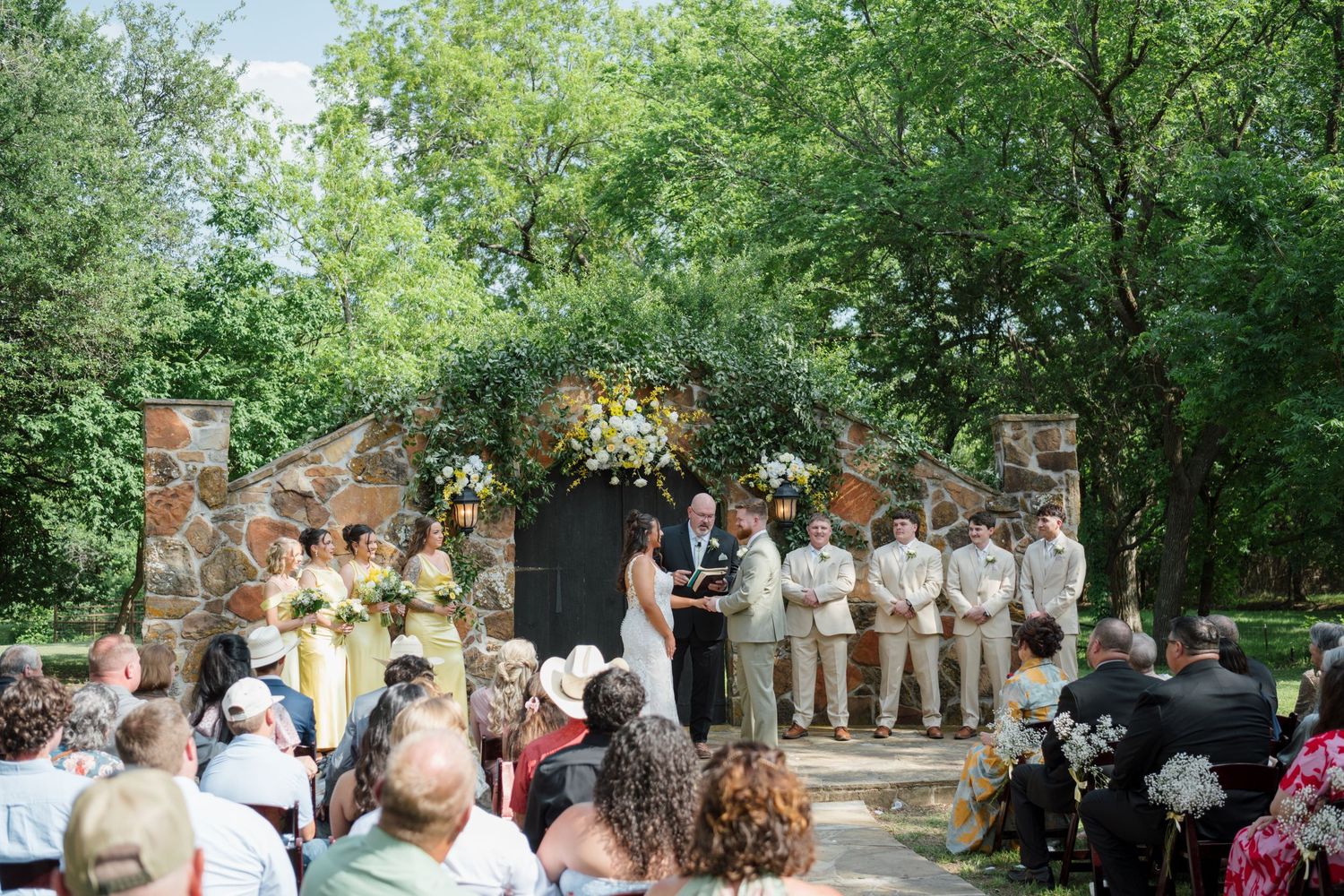 Outdoor wedding ceremony in a lush garden with stone arch, floral arrangements, wedding party, and seated guests.