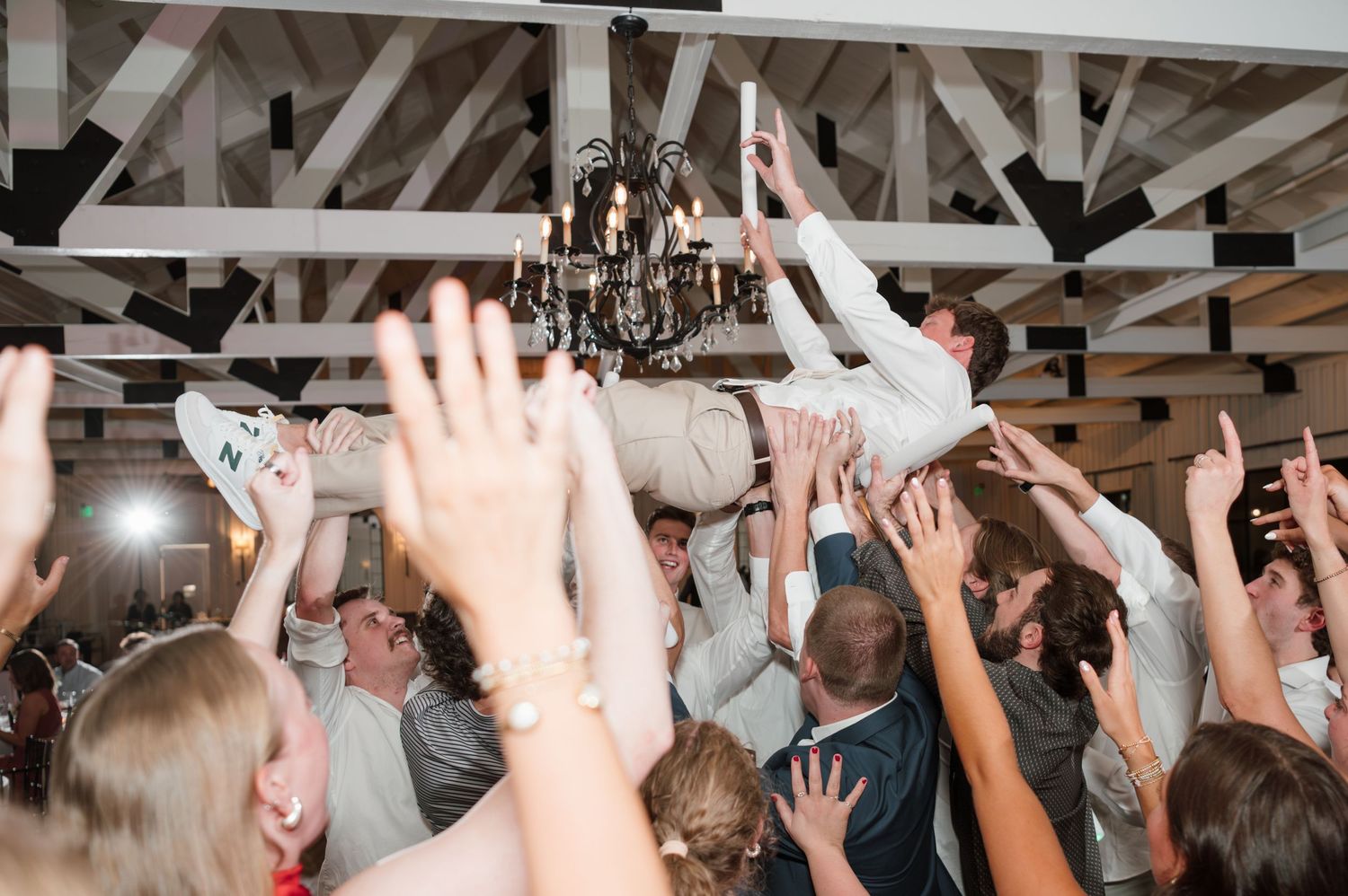 Wedding guests raise hands in celebration during reception under rustic wooden beams.