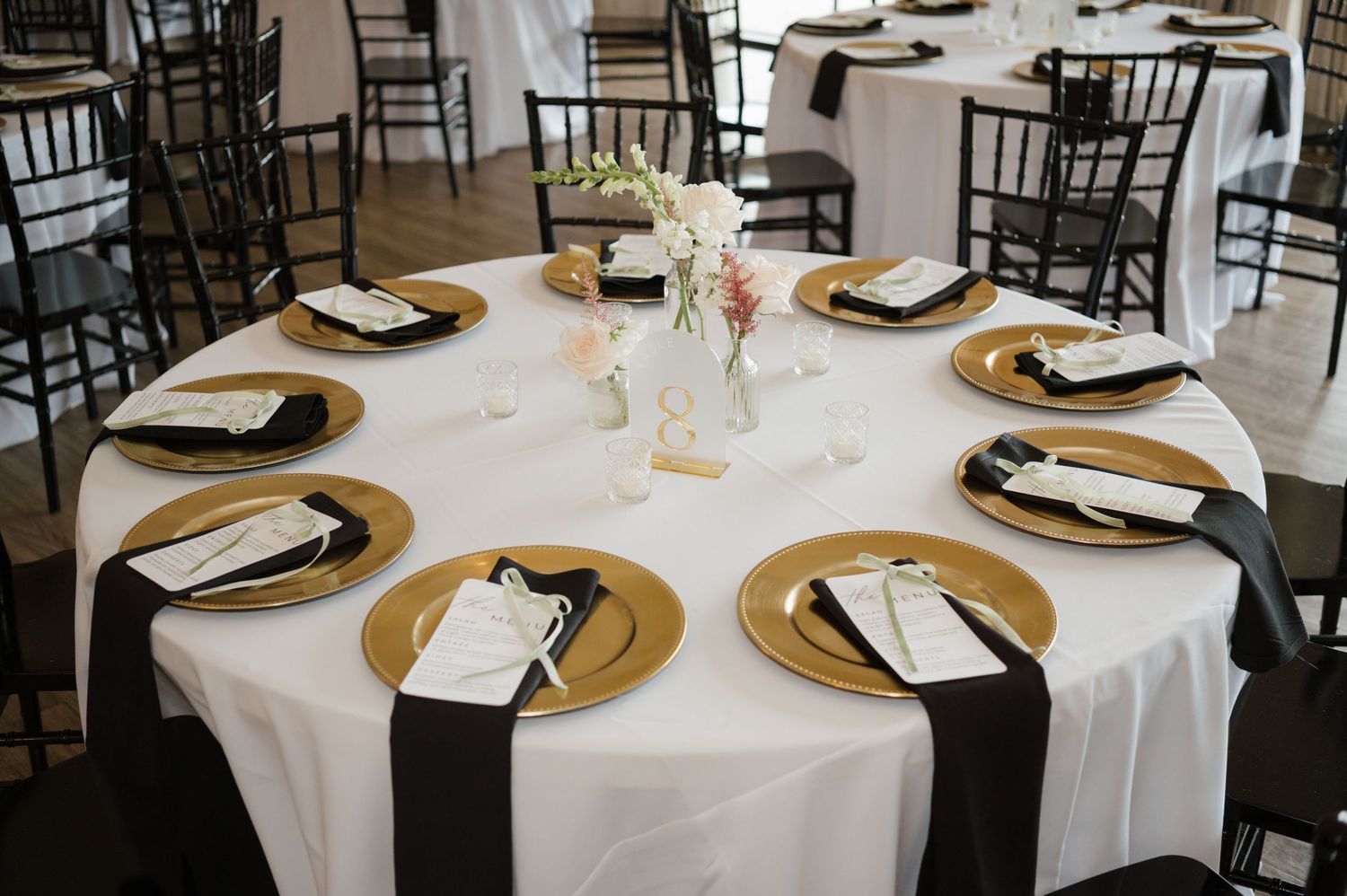 Elegant wedding reception tables set with gold chargers, white linens, and black chairs in a bright indoor venue.