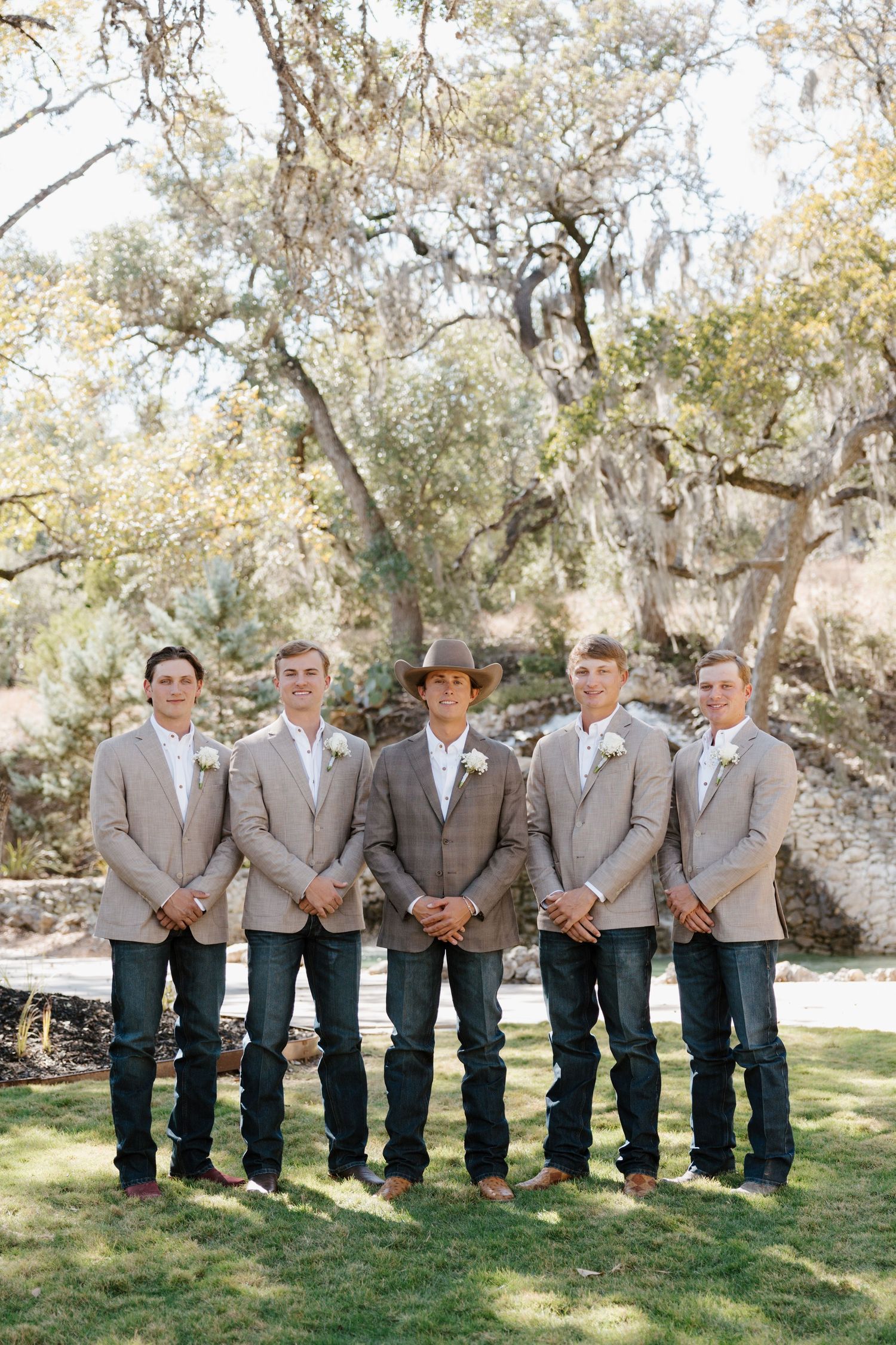 Groomsmen in matching gray jackets and jeans pose together on grass under oak trees.