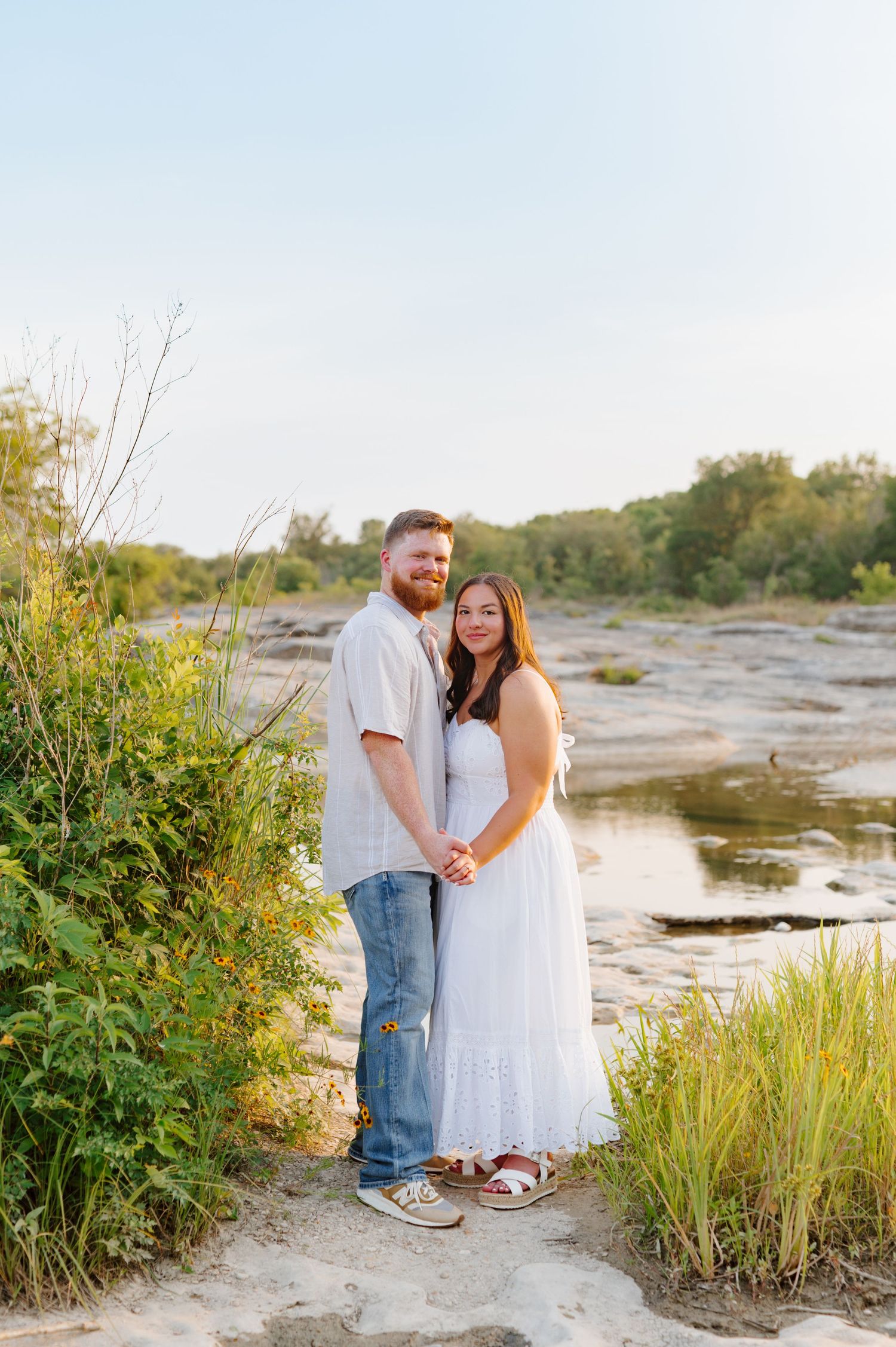 A couple shares a romantic moment on a sandy beach path surrounded by coastal vegetation at sunset.