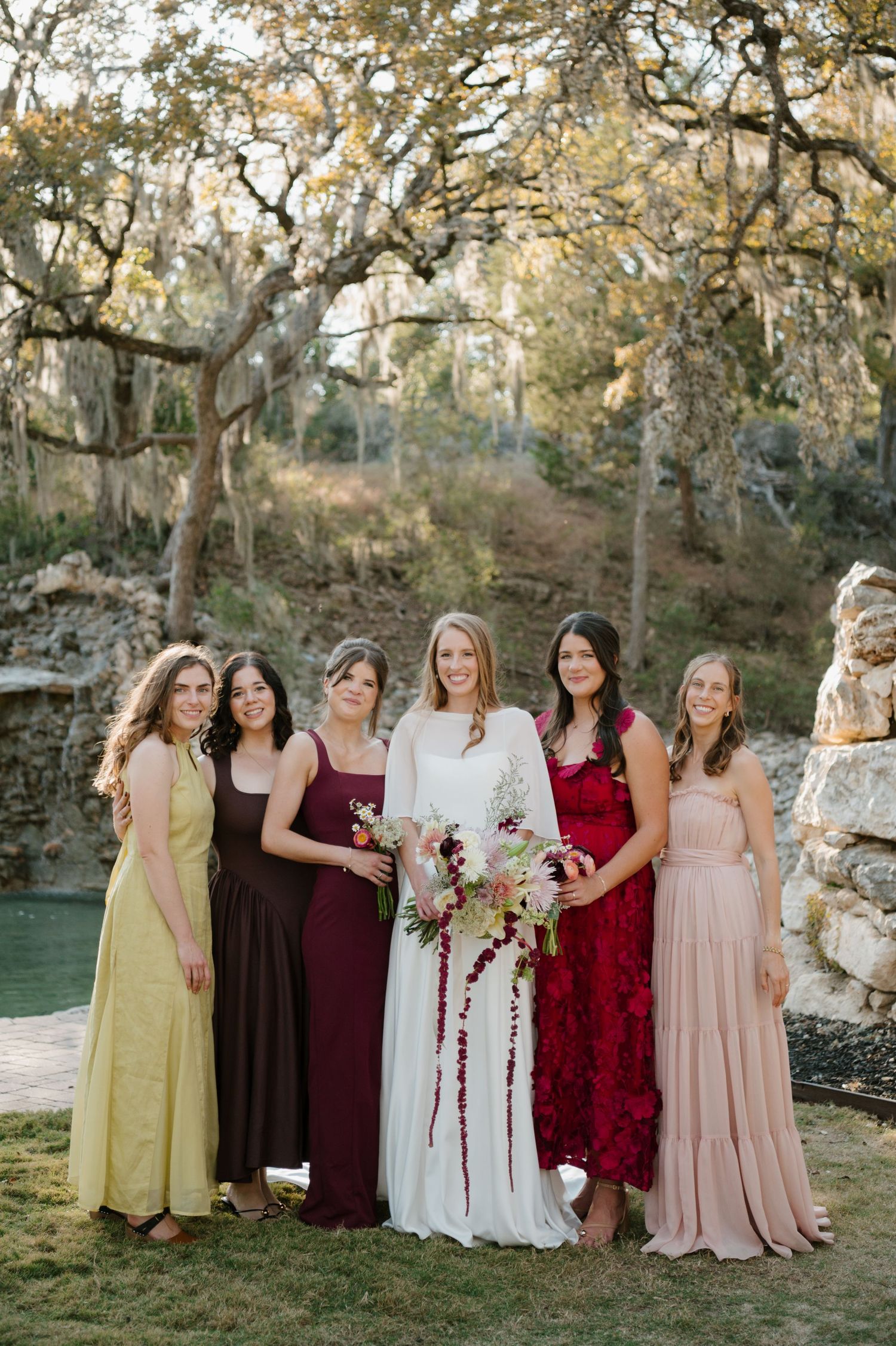 Wedding party in mix of burgundy, pink and brown dresses standing together outdoors.